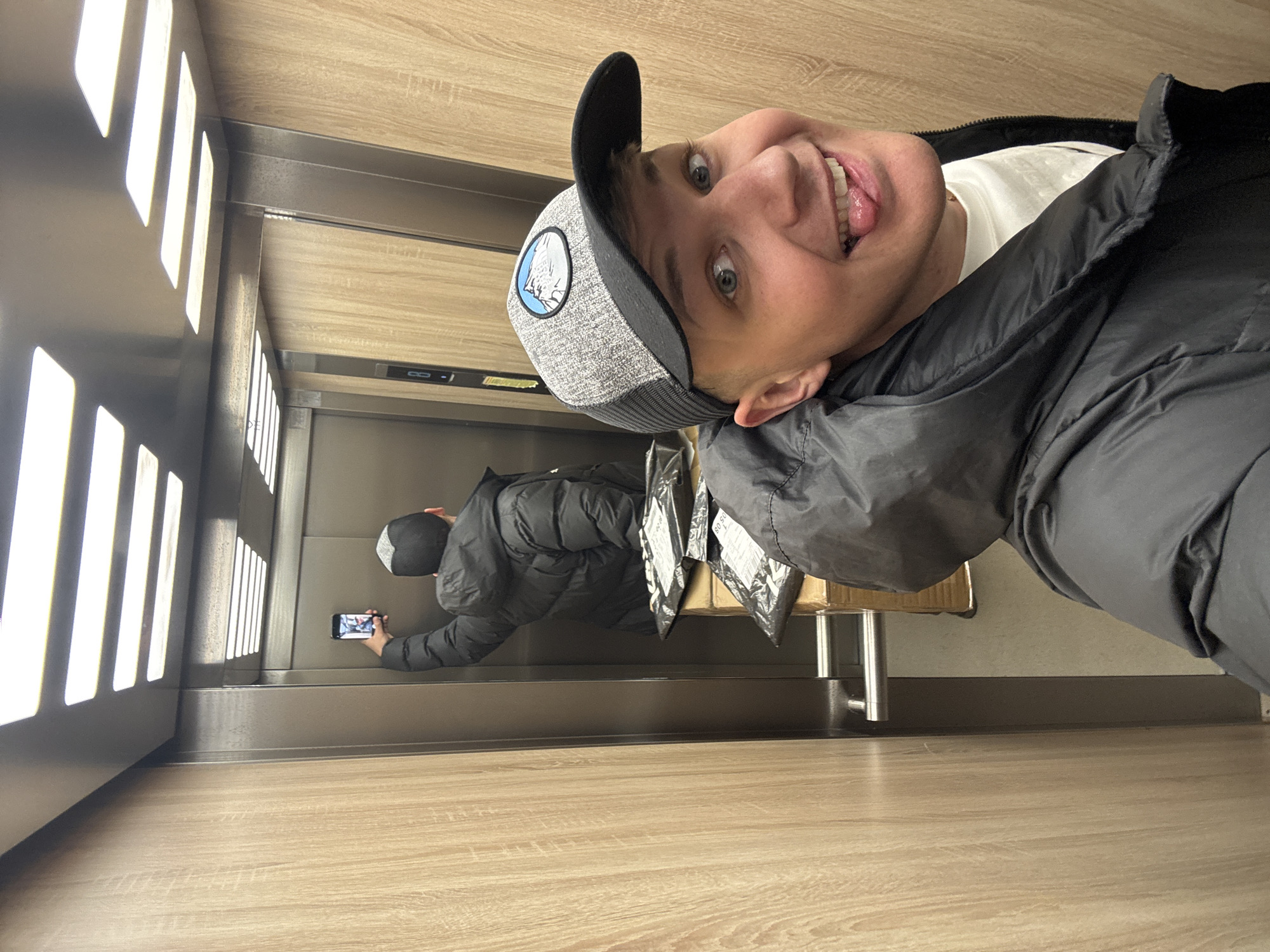 A man with a black jacket and gray baseball cap is taking a selfie in a mirrored elevator with packages on a stool. The location appears to be an elevator; the building and city cannot be determined from the image.