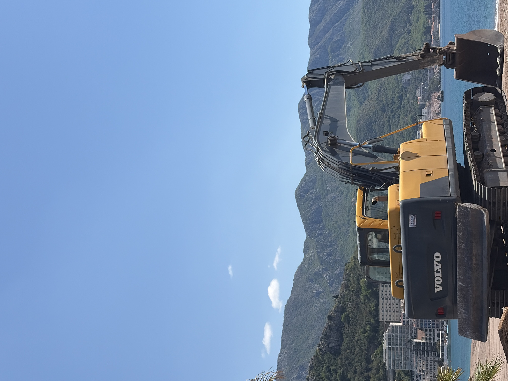 A Volvo excavator is positioned with mountains, the city of Bečići, Montenegro, and the sea as the backdrop. The excavator is predominantly yellow and grey, with the Volvo logo visible on the rear. The excavator's arm and bucket are extended. The location appears to be an elevated position overlooking Bečići, with buildings visible amongst the green mountains leading down to the coastline. The sky is clear and blue.