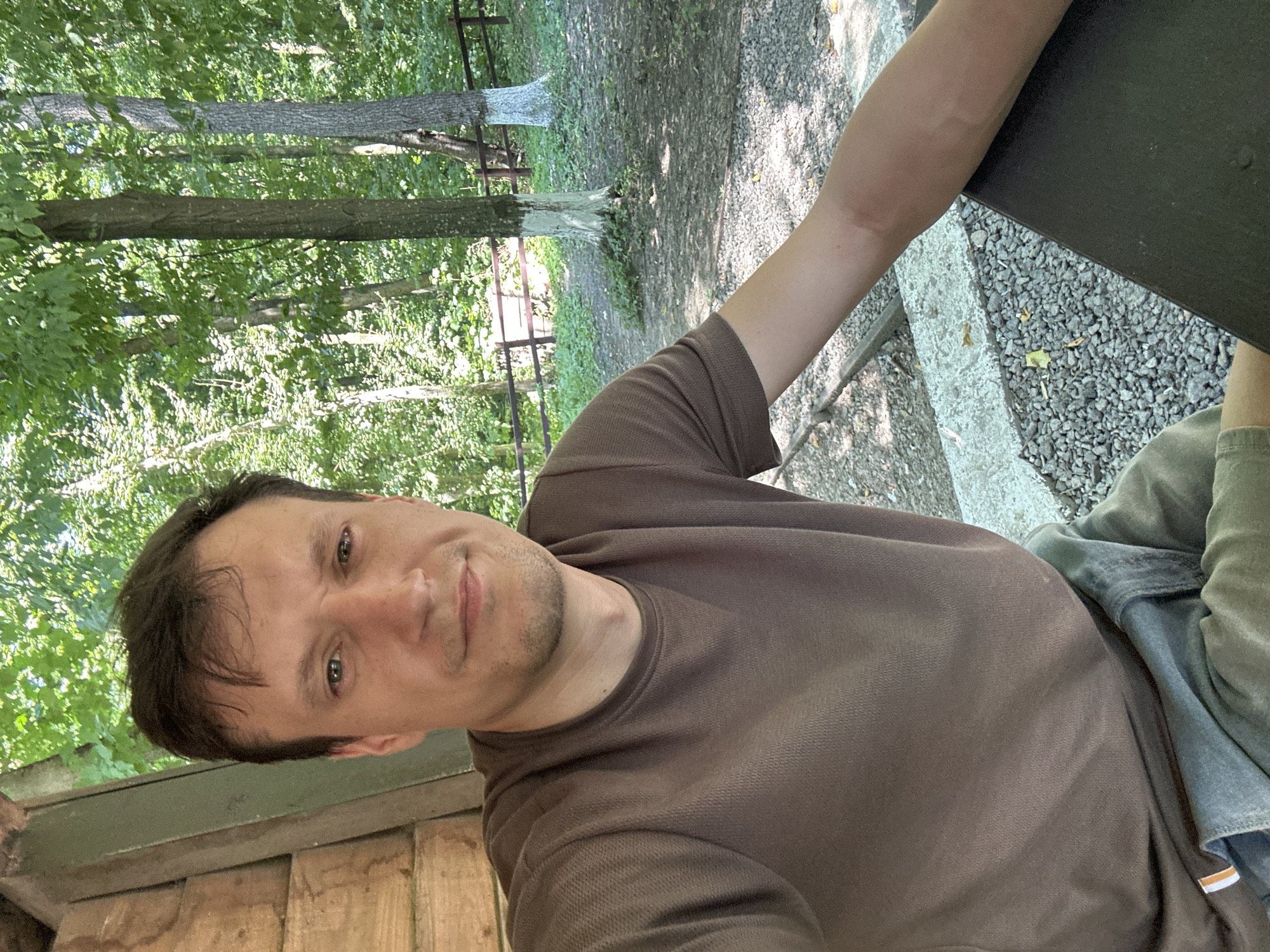 The image is a selfie taken outdoors, likely in a park or wooded area, with a focus on the person and the natural environment. The subject, a male with light skin and brown hair, is positioned in the foreground, facing the camera with a neutral expression. He is wearing a brown t-shirt and appears to be seated at a table.
The background reveals a lush green forest with tall trees. Some trees have white paint on their lower trunks, a common practice for protection or identification in certain regions. A wooden fence runs horizontally through the mid-ground, separating the immediate area from the deeper woods. The ground is covered with small stones and gravel, with patches of dirt and fallen leaves. The overall lighting suggests a sunny day.
The location is identified as Shatava, Ukraine, a place known for its natural beauty and likely a recreational area. The scene depicts a person enjoying a moment outdoors, possibly during a casual outing or visit to the area.