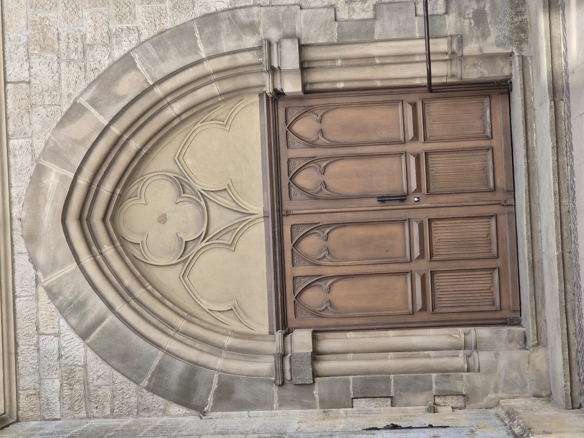 The image shows a close-up, eye-level view of a large, ornate wooden double door set within a Gothic-style stone archway. The doorway is flanked by carved stone pillars and is accessed by a set of stone steps. The doors themselves are a rich brown wood with decorative paneling, featuring pointed arch motifs carved into the upper sections. The stone archway is made of light-colored, weathered stone blocks and features multiple concentric curves. Above the doors, within the arch, are decorative stone carvings. The surrounding wall is constructed of rough-hewn, light-colored stone blocks. A dark metal handrail is visible to the right of the doorway. The scene appears to be an entrance to a historic building. This is the entrance to the Saint-Michel church in Differdange, Luxembourg.