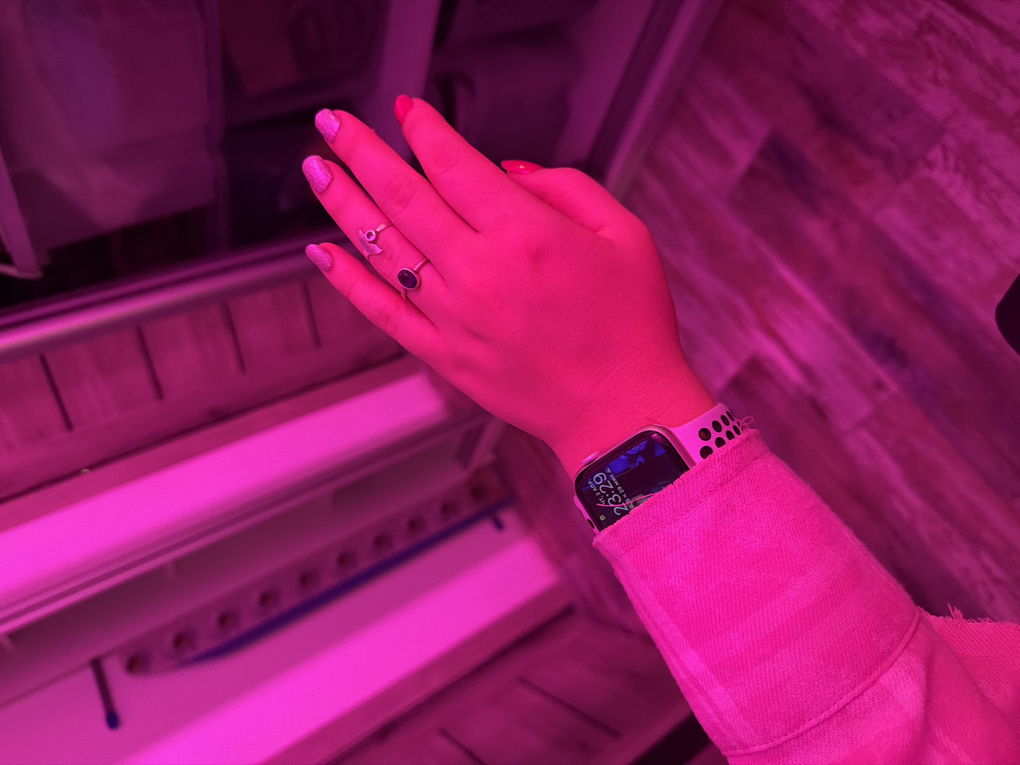 A right hand and forearm are visible under pink lighting. The fingers are adorned with pink glitter and solid pink nail polish, as well as two silver rings. An Apple Watch with a pink strap is on the wrist, displaying a watch face with a time of 23:29. The arm is covered by a pink jacket or shirt. The background includes what seems to be part of a storage system with horizontal white supports. The scene appears to be indoors, possibly in Большая Шильна, Russia.