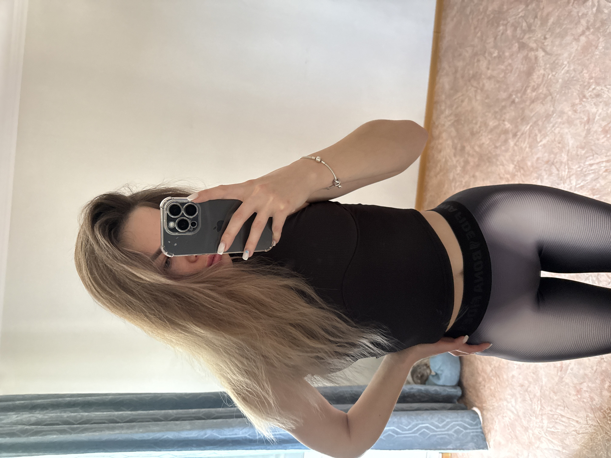 An indoor mirror selfie depicts a person, likely female, captured from the waist up. The image is rotated 90 degrees counter-clockwise, showing the person's head on the left and body extending to the right. They have long, light brown hair that flows over their shoulder and partially covers their face.
The person is wearing a black ribbed crop top and tight-fitting dark grey leggings with a subtle horizontal striped pattern. A silver bracelet with small charms adorns their right wrist. They hold a smartphone with a clear case and a prominent camera module, obscuring part of their face; only their eyes and a portion of their lips are visible behind the phone and hair. One hand rests on their hip, pressing slightly into the waistband of the leggings.
The background, visible in the mirror's reflection, includes a plain light-colored wall on the upper left and a textured, possibly wallpapered wall with a reddish-brown hue on the right. At the bottom of the frame, blue-grey curtains and a light blue upholstered object are visible. The lighting is bright and appears to be natural daylight. The white text on the elastic waistband of the leggings is partially visible and reversed, appearing to read "DEADBONES".