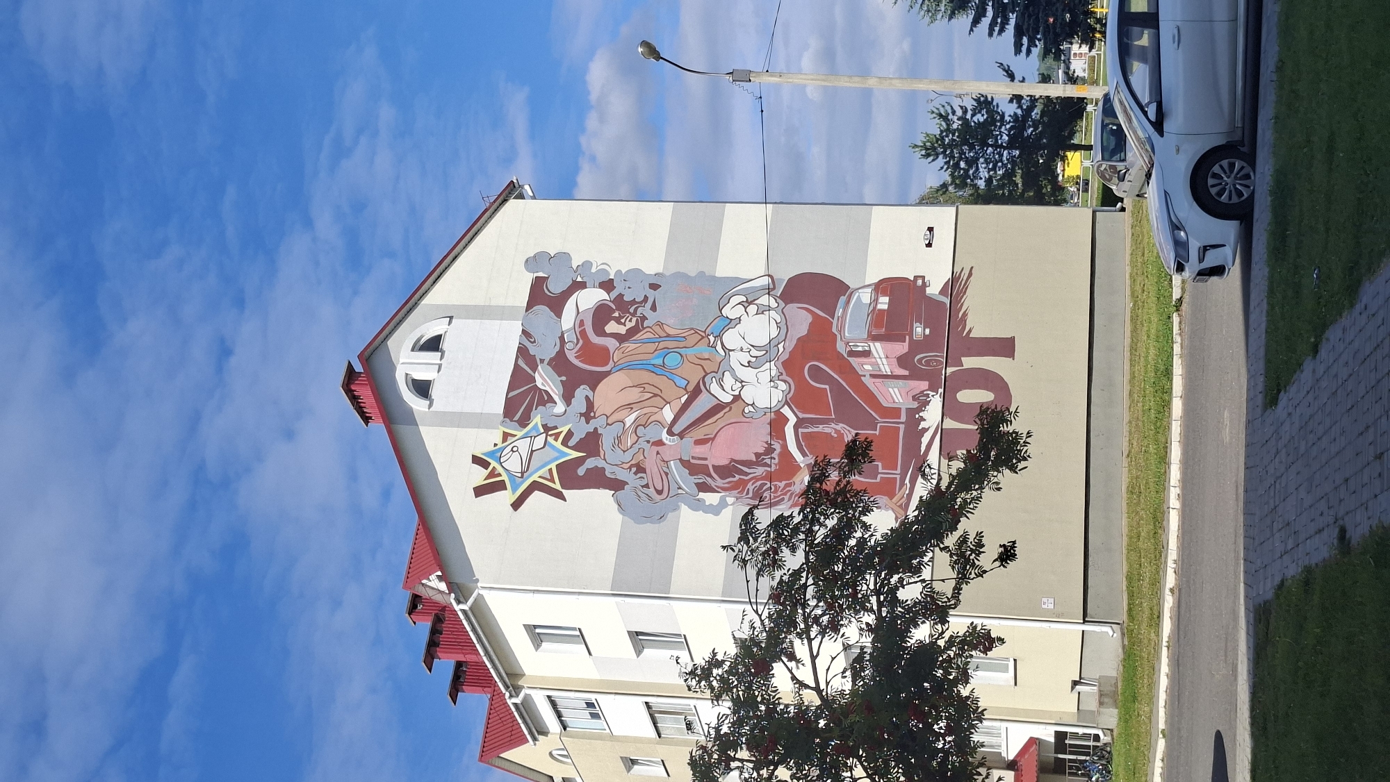 A mural depicting a firefighter is painted on the side of a multi-story apartment building in Myadzyel, Belarus. The mural features a firefighter in full gear, holding a hose and surrounded by smoke. A red fire truck and the number "112" are also visible in the mural. The building has a red roof and light-colored walls with windows. In the foreground, there is a tree with red berries, a paved walkway, a patch of grass, and a street with a white car parked on the side. A lamppost stands near the building. The sky is blue with scattered clouds.