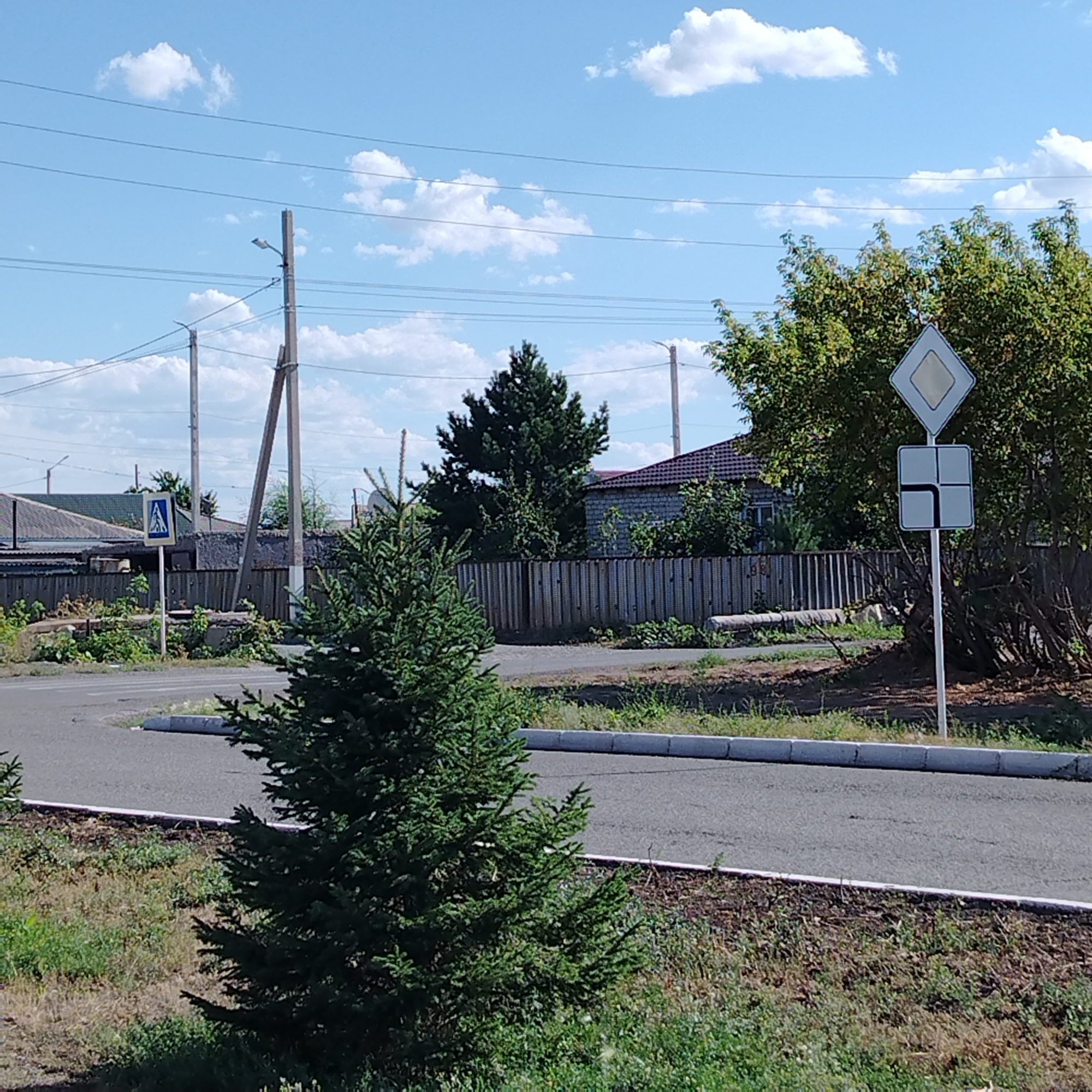 The image depicts an outdoor scene in Ekibastuz, Kazakhstan, on a clear day with scattered clouds. A road is visible in the foreground, with a small evergreen tree to the left. In the mid-ground, a wooden fence runs horizontally, separating the road from a residential area. Beyond the fence, houses with tiled roofs and brick walls are visible. Electrical poles and power lines stretch across the sky. A pedestrian crossing sign is posted on a pole near the road, and a diamond-shaped sign with a white interior is mounted on another pole. The environment appears to be a semi-urban or suburban setting. There are no people or animals visible in the image, nor are there any specific actions or interactions taking place. The overall impression is one of a quiet, ordinary day in a residential area.