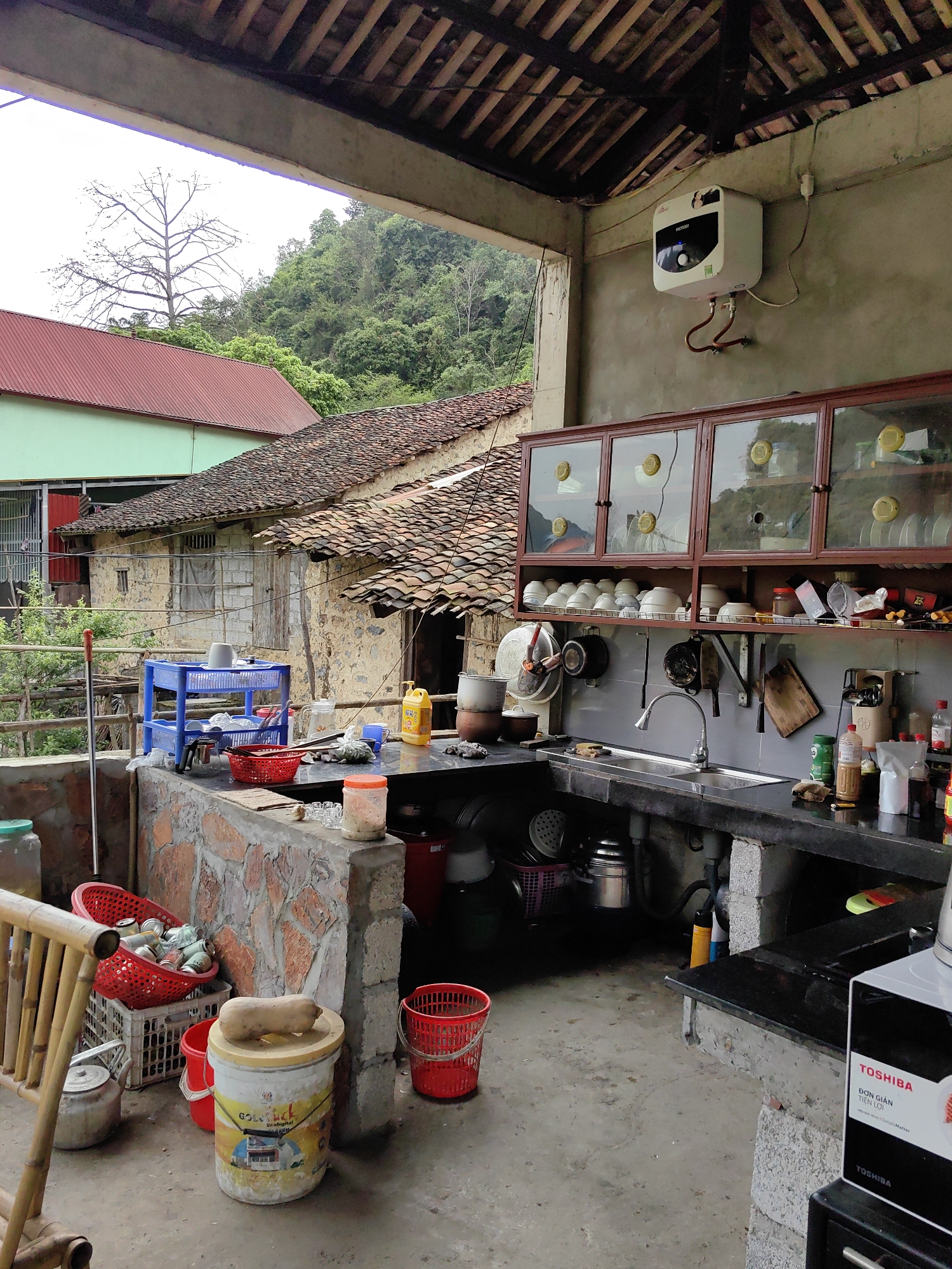 This outdoor kitchen is located in Xã Đàm Thủy, Vietnam. It features a sink, countertop, and cabinets stocked with dishes and utensils. An electric water heater is mounted on the wall. Outside the kitchen area, there are other buildings and lush green vegetation in the background. Red buckets and other storage containers are scattered around the floor.