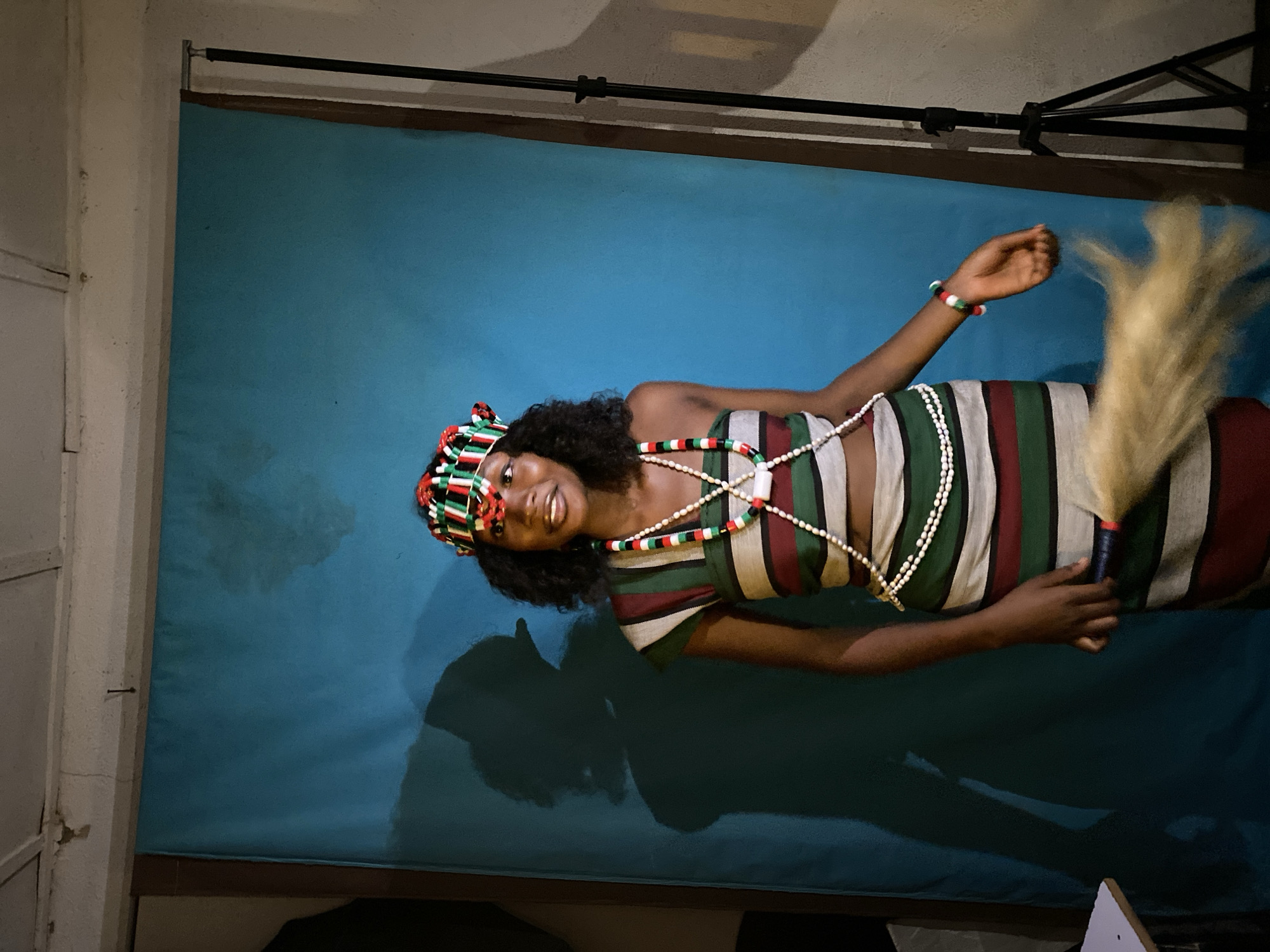 The image depicts a woman indoors in Durimi, Nigeria, against a solid blue backdrop likely used for studio photography. She is wearing traditional Igbo attire, including a horizontally striped dress in white, green, and burgundy colors, and beaded necklaces and headwear in red, green, white, and black colors. She's holding a traditional horsetail fly whisk in her right hand. Lighting equipment is visible in the background, including a support stand and what appears to be a softbox or light. The photograph may have been taken in a photo studio or a prepared indoor setting.