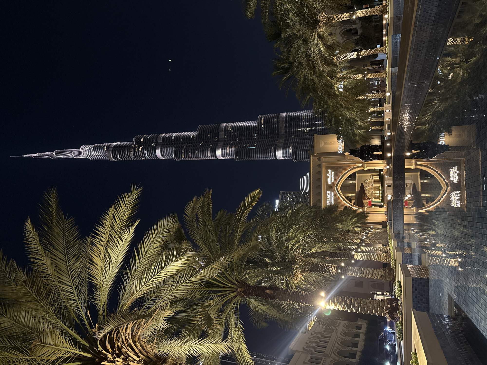 The image shows the Burj Khalifa at night, viewed from an outdoor space in Dubai, UAE. The skyscraper is illuminated and positioned vertically on the left. Palm trees frame the bottom and right of the scene. A walkway with string lights is on the right, next to a tiled fountain reflecting the nearby buildings and trees. Several people are discernible as blurry figures in dark clothing near the fountain. The sky is dark blue.