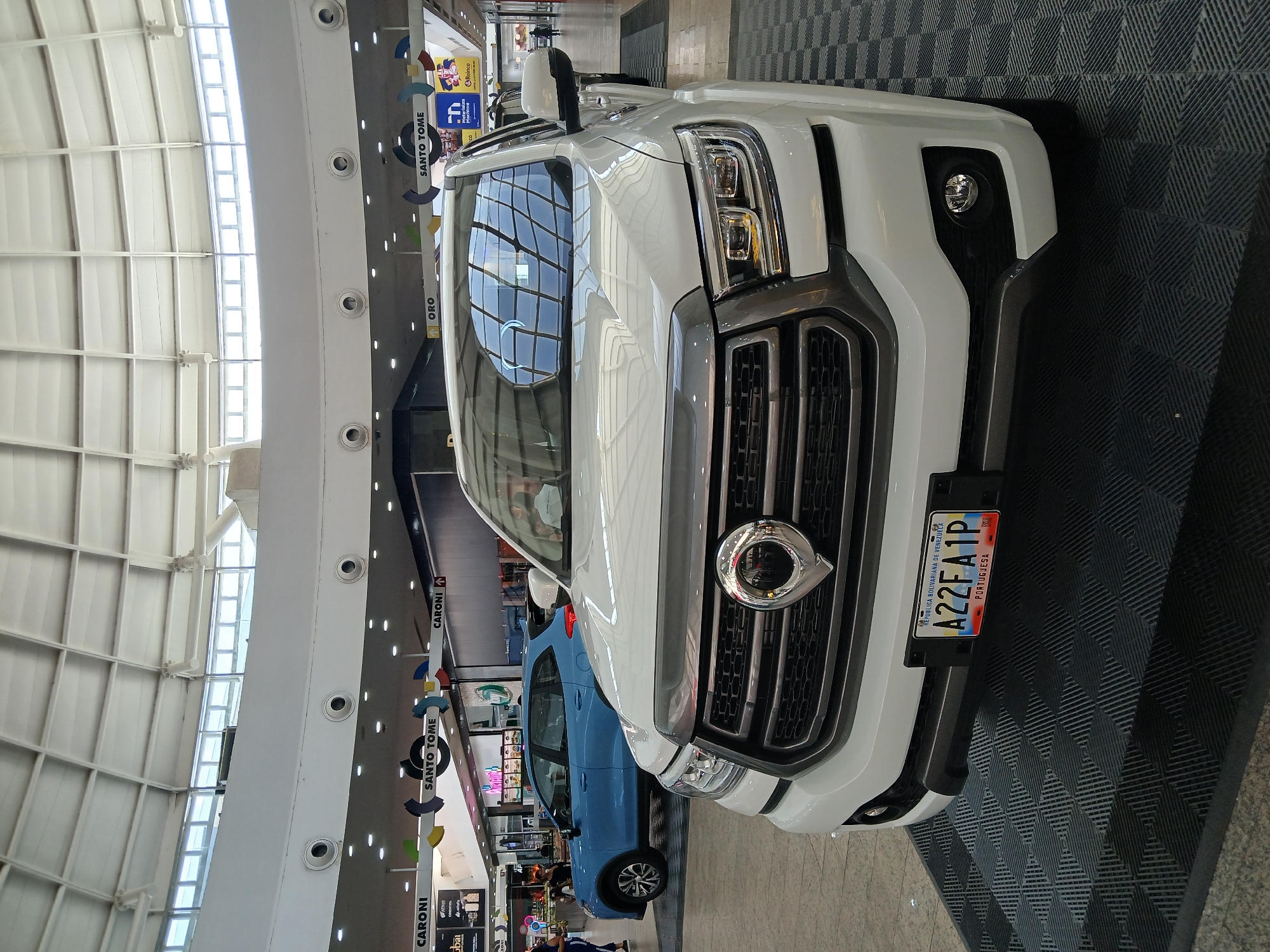 The image shows a white Great Wall Wingle 7 pickup truck, displaying the license plate "A22FA1P" from the state of Portuguesa, Venezuela. The truck is parked indoors, possibly at a dealership or an event inside a shopping mall. The background shows retail signs for "CARONI" and "SANTO TOME," suggesting the location is within the Ciudad Guayana area, Venezuela. Another blue car is visible behind the truck. The floor has a dark, geometric pattern. The building has a high ceiling with a skylight design.