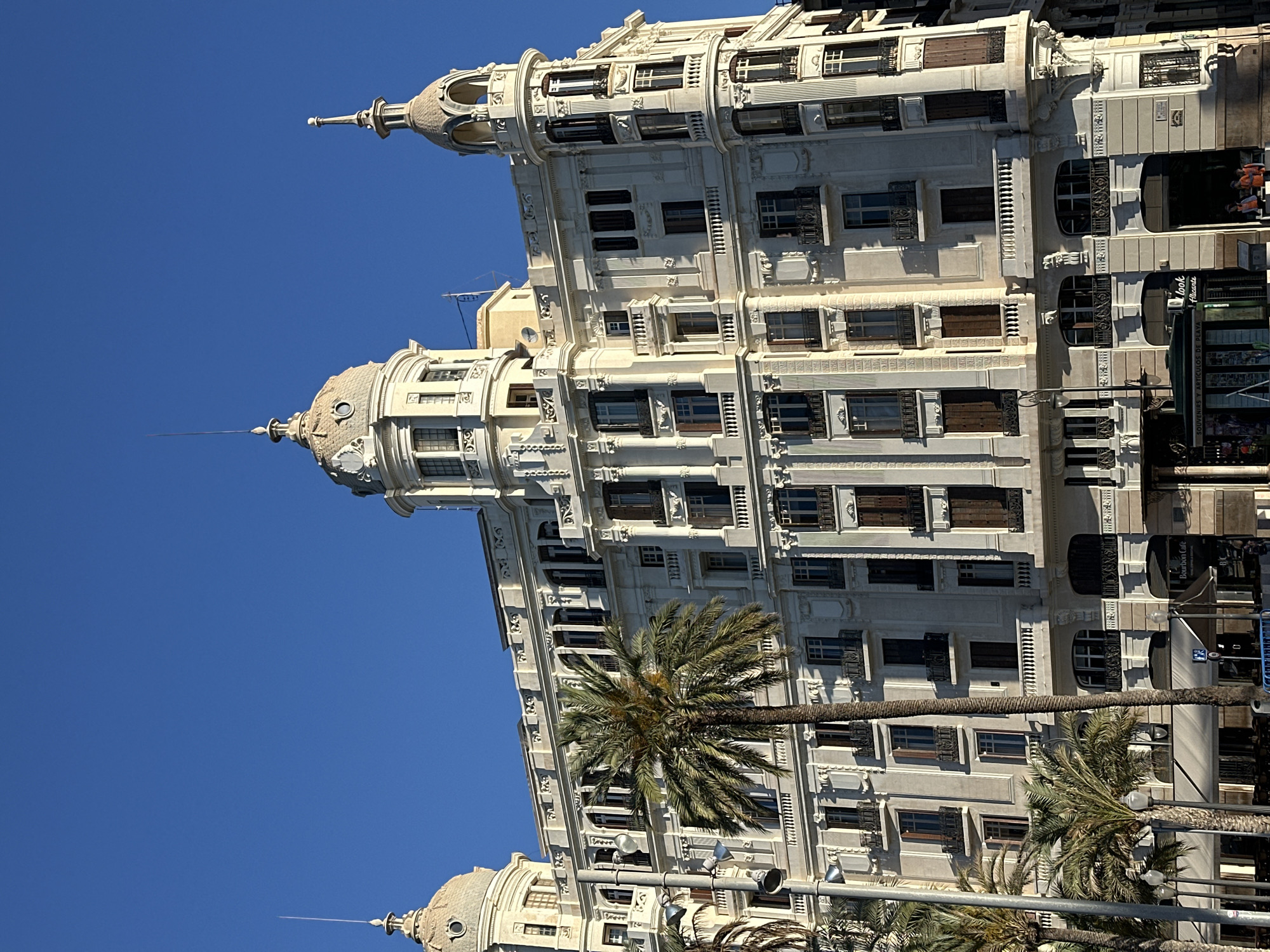 The image shows a tall, ornate, light-colored building under a clear blue sky. The building has multiple stories with rows of windows, and decorative architectural elements on the facade. Palm trees are visible in front of the building. Signs and other urban elements suggest the building is located in a city.