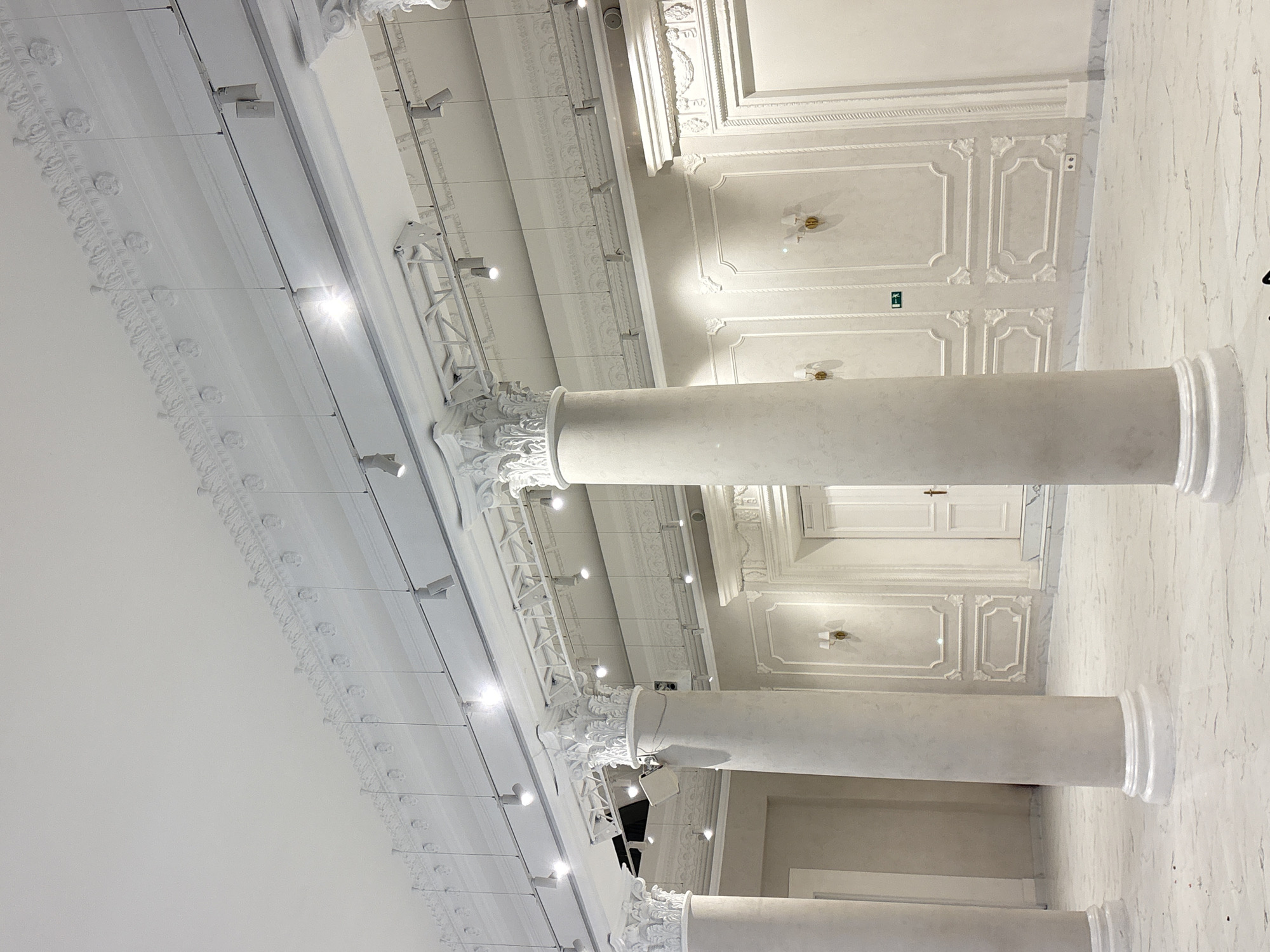 The image shows the interior of the Perm Academic Theater, located in Perm, Russia. The perspective is low, looking upward toward the ceiling and the columns that support it. The walls and ceiling are predominantly white with ornate, classical-style decorative moldings. The lighting consists of numerous small spotlights suspended from the ceiling, along with sconces on the walls. Marble or marble-like finish on the wall. No people are visible in the image.