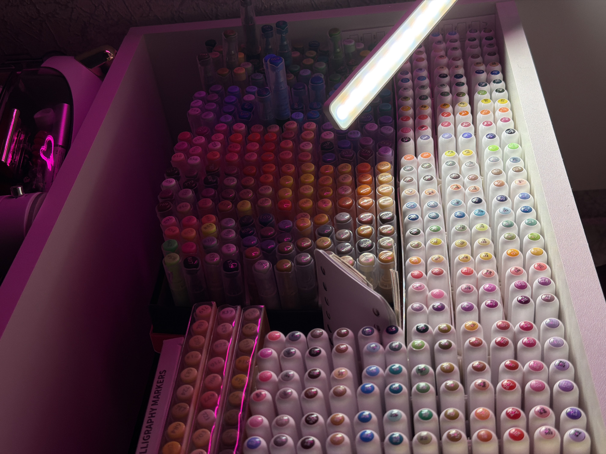 The image shows a shelf filled with art markers. The markers are organized in clear plastic holders, arranged in rows and columns. The majority of the markers are white with colored caps or tips, indicating different colors. Some markers have visible numerical codes on them. To the left, a pink label reads "LLIGRAPHY MARKERS". Above the shelf, a pink light illuminates the top of the image. A bright LED lamp is positioned in front of the markers on the right side of the shelf, casting a glare. The background is dark and textured, suggesting a wall. The overall impression is of a well-organized collection of art supplies.