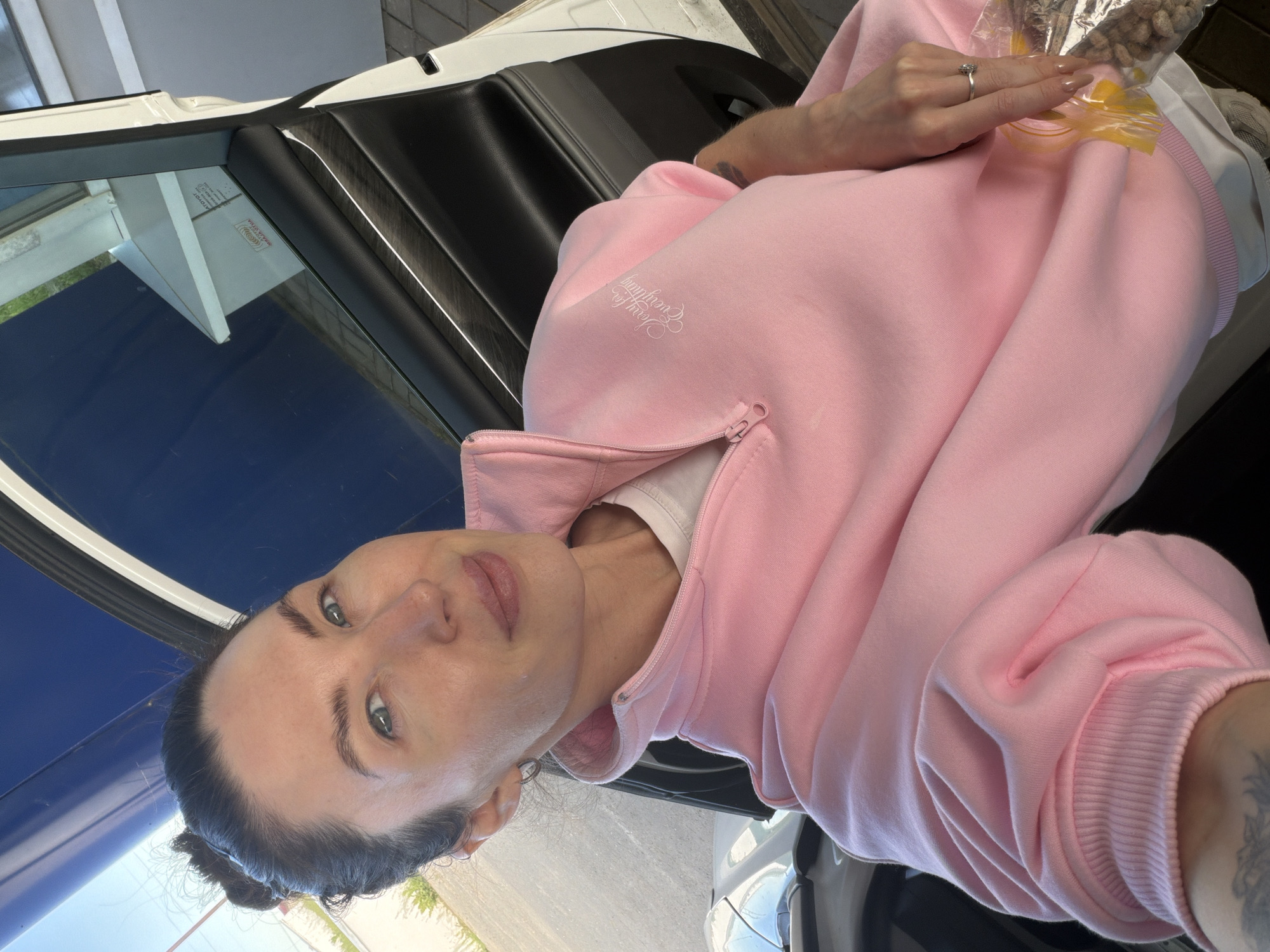 A woman is captured in a well-lit, candid shot, looking directly at the camera with a neutral expression. She has dark hair pulled back from her face, and her eyes are light in color. She is wearing a light pink zippered sweatshirt over a white undershirt. The sweatshirt features stylized white text on the chest that appears to read "Love for Everything." Ribbed cuffs are visible on the sleeves.
She is seated in what appears to be the passenger seat of a car, with the car door open. The interior features black door panels and a dark dashboard. Her left hand, adorned with a silver ring on the ring finger and light-colored nail polish, holds a clear plastic bag containing light-colored, possibly crunchy snacks like nuts or cereal. A yellow elastic or tie is securing the bag. A tattoo is partially visible on her left arm.
The setting appears to be outdoors during bright daylight, suggested by the clear blue sky and strong light visible through the car windows and reflected on surfaces. Reflections in the car window show hints of an outdoor environment, including some greenery, paved ground, and a building or structure. Partially visible text in the reflection includes "AVTOTOY" and "NORDIC HOUSE" (reversed), possibly indicating businesses or markings in the vicinity. The scene takes place in Ильинское, Russia.