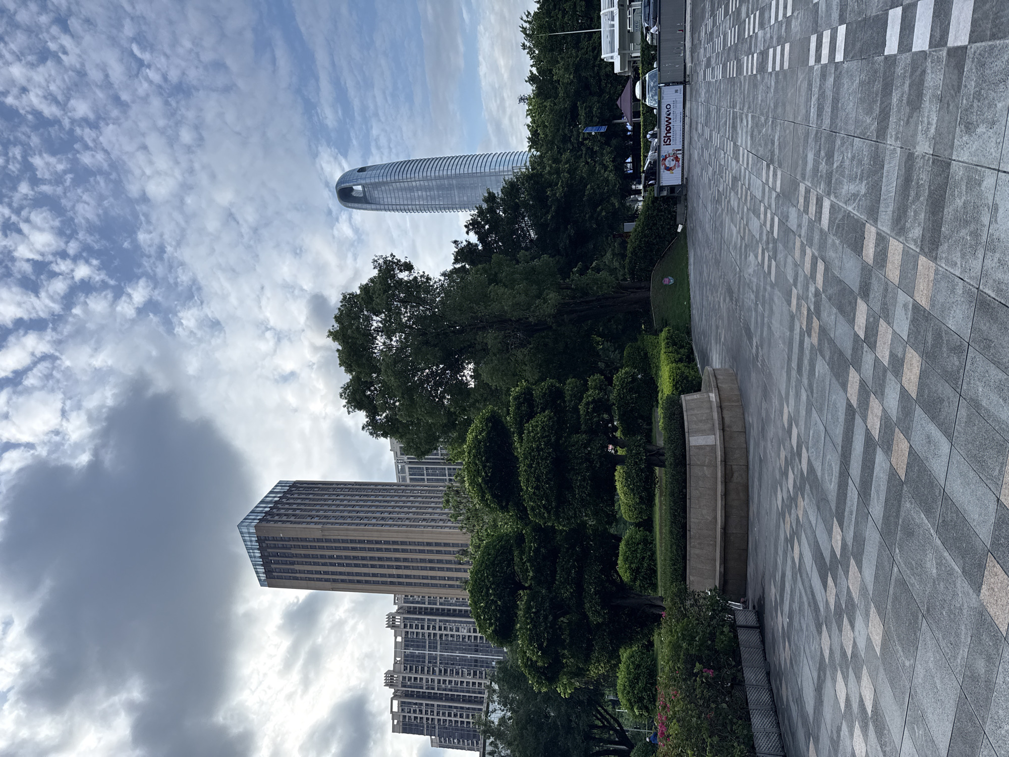 This is a medium-angle, eye-level shot of a public plaza in Haizhu District, China. In the foreground, a patterned tiled plaza with a light and dark grey checkerboard design stretches out. To the left, a raised planter bed with manicured green bushes and a circular stone seating area is visible. Two topiary trees with rounded canopies stand within the planter. Further back on the left, several residential apartment buildings are visible. Dominating the background are three distinct skyscrapers. The tallest and most prominent is a modern, curved glass skyscraper with a distinctive oval opening near its top, located on the right side of the frame. To its left and slightly closer to the viewer is a rectangular, beige skyscraper topped with glass. Behind the residential buildings and to the left of the beige skyscraper is another tall residential building. Lush green trees and foliage fill the middle ground between the plaza and the skyscrapers. The sky above is a mix of grey clouds and patches of blue sky with scattered white clouds. Several cars and a white tent structure are visible behind a low barrier at the far right of the frame, along with a banner that reads "iShow2.0". A few individuals are faintly visible in the background, one near the planter on the left and others near the barrier and vehicles on the right.