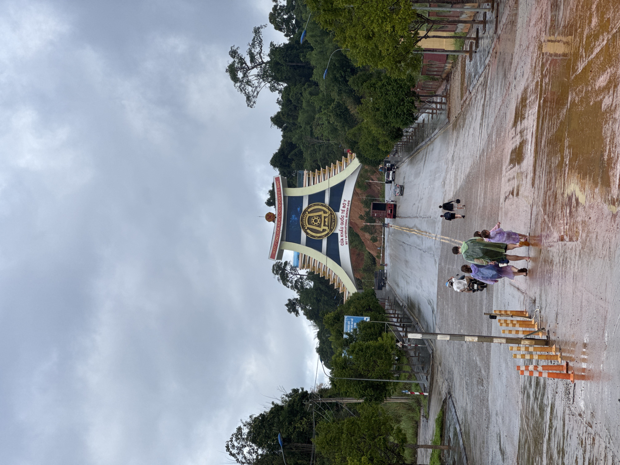 The image presents a wide view of the Bờ Y International Border Gate in Vietnam on an overcast, damp day. A large, ornate archway serves as the central feature, spanning the road and marking the entrance or exit. The sky above is heavily clouded, and the asphalt road is visibly wet with puddles and reflections, suggesting recent rainfall or ongoing light rain.
Several people are seen walking on the wet road, some wearing rain ponchos in shades of purple and green, indicating the rainy conditions. A few vehicles, including cars and a truck, are also present on the road. Lush green trees and hills flank the road and rise behind the archway, providing a natural backdrop. On the right side of the road, orange and white traffic cones and barriers are visible. A utility pole with wires stands closer to the foreground on the right.
The archway prominently displays text in both Vietnamese and English. Visible text includes: "NƯỚC CỘNG HÒA XÃ HỘI CHỦ NGHĨA VIỆT NAM" (Socialist Republic of Vietnam), "KON TUM", and "CỬA KHẨU QUỐC TẾ BỜ Y - BƠ Y INTERNATIONAL BORDER GATE". The scene captures a moment of daily activity at the border post under subdued weather conditions.