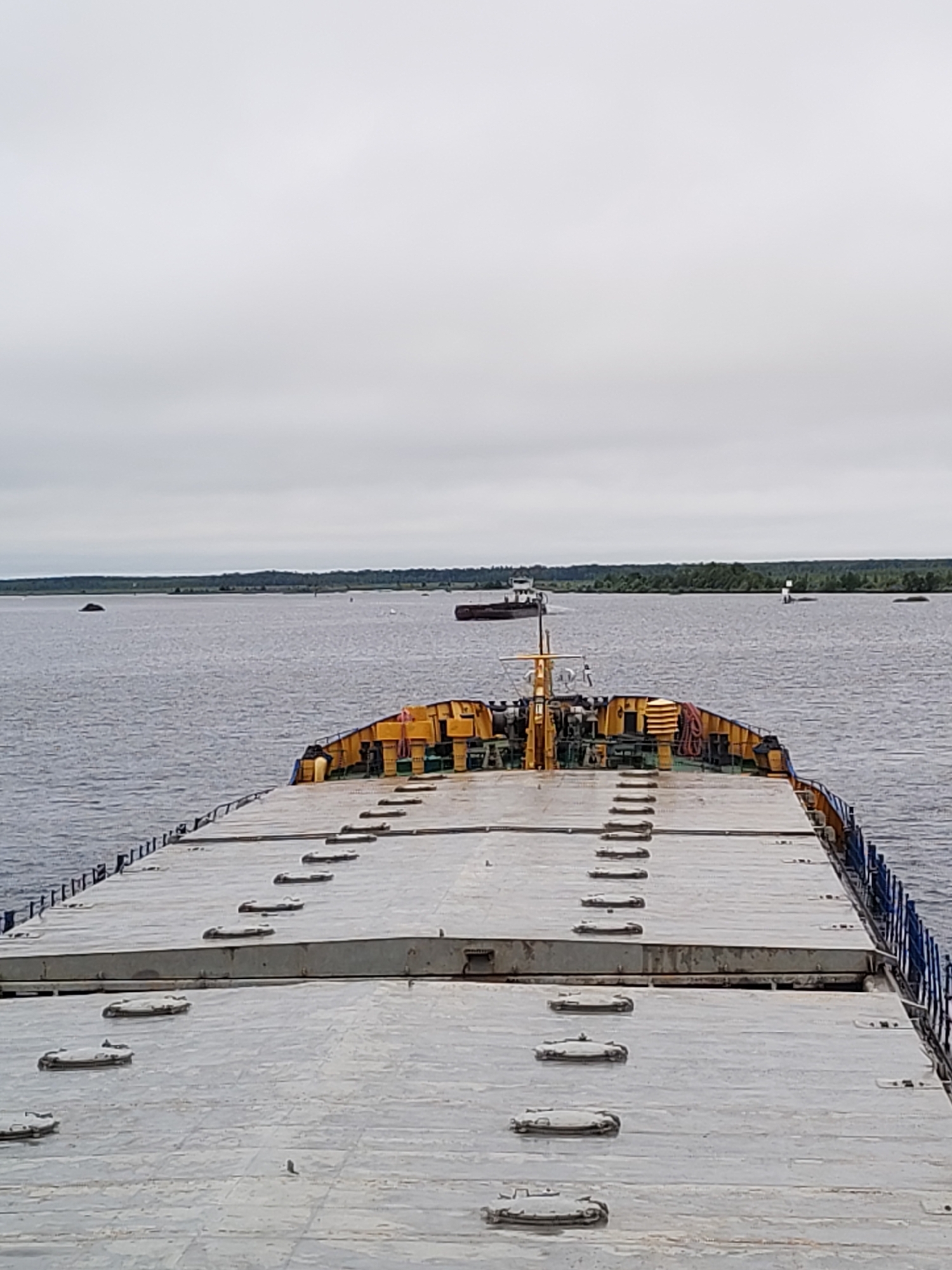 The image shows a view from the deck of a large, flat-topped barge or ship, looking forward and slightly across a body of water under a cloudy sky. The foreground is dominated by the textured, grey deck of the barge, featuring numerous circular or oval hatches with raised rims. These hatches are arranged in rows. Further back, the barge has a raised structure with yellow and green elements, including railings and what appears to be equipment or cargo. A mast with rigging is visible in the center of this structure.
In the water, several other vessels are present. A smaller, darker vessel is moving away in the distance towards the left. To the right of center, a white buoy or navigational marker stands in the water, with another dark vessel, possibly a barge, further beyond it. The background consists of a distant shoreline with a dense green tree line, suggesting a forested area. Several dark, irregular shapes, potentially small islands or large rocks, are scattered in the water near the shoreline.
The location is identified as Novokemskii, Russia. The scene depicts a calm, overcast day on a waterway, with industrial or cargo vessels in transit or at anchor. There are no people visible in the image. The overall impression is one of a working waterway environment.