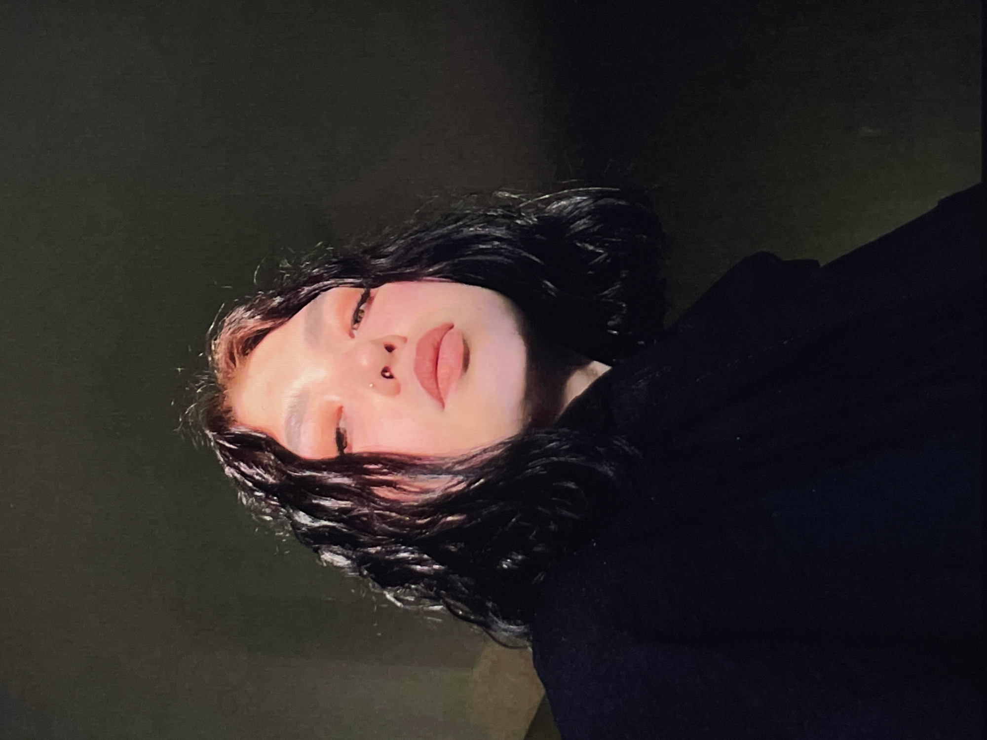 A person with dark, wavy hair and light skin is depicted in a close-up shot with a dark background. The person has their eyes partially closed and a piercing on their septum. They are wearing a black garment, and the composition is such that the person's face is centrally positioned. The location is not specified.