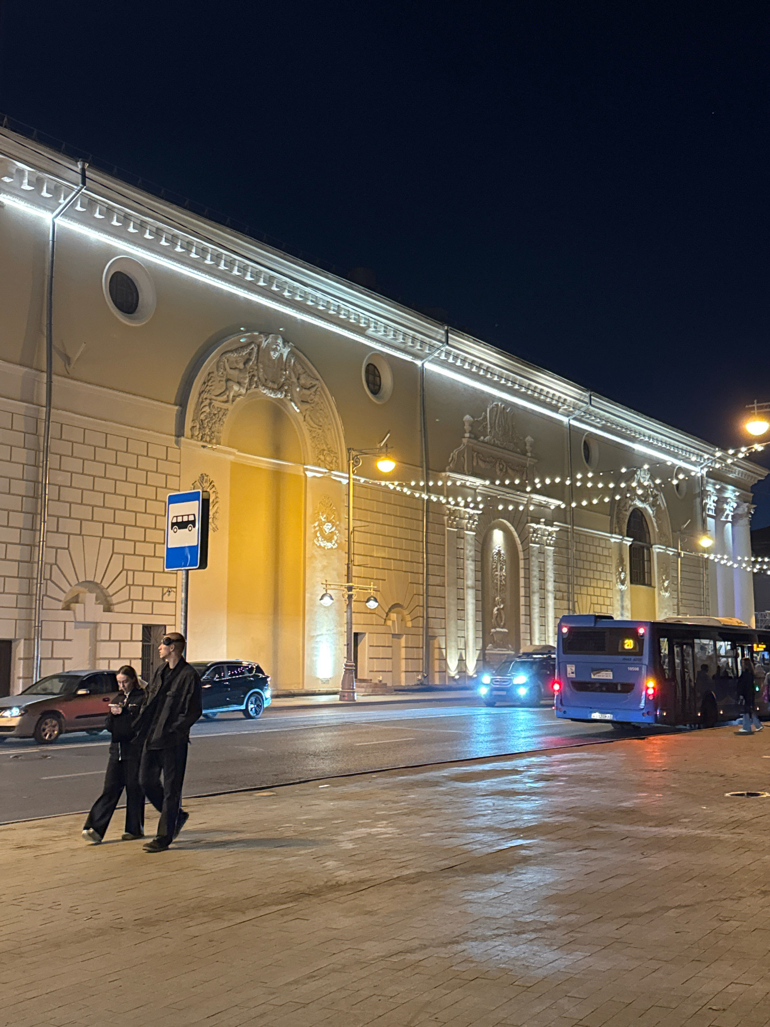 The image depicts a night scene on a city street, possibly in Tver, Russia, near a large building with ornate architectural details and illuminated by strings of lights. In the foreground, two individuals, a man and a woman, are walking side-by-side on a paved sidewalk. The man is taller and appears to be wearing dark clothing, including pants and a jacket, and has what looks like goggles pushed up on his forehead. The woman is shorter and is also dressed in dark, loose-fitting clothing. They are walking towards the left of the frame.
To their right, a blue bus is stopped at what appears to be a bus stop, indicated by a blue and white sign with a bus icon. The bus has its doors open, and several passengers are visible inside. The bus is facing towards the right of the frame. There are also cars visible on the street, including a dark SUV with its headlights on and a red sedan parked further down the road. The street is wet, reflecting the lights from the surrounding environment. The building in the background is multi-story, with arched windows and decorative elements. The sky is dark, indicating nighttime.