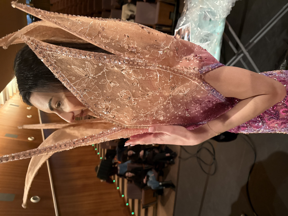 An individual is depicted in profile, facing left, within an auditorium in Paris, France, likely a grand amphitheater found in a prominent educational or cultural institution like the Sorbonne University. The subject is a woman with dark hair and fair skin, wearing an elaborate, two-toned garment. The upper portion of the garment consists of sheer, light peach-pink lace adorned with intricate floral embroidery and iridescent sequins, forming stiff, petal-like structures around her neck and shoulders. This transitions into a densely sequined lower section in fuchsia and purple hues. Her right hand, with manicured light pink nails and a thin gold bracelet, is positioned against the garment near her chest.
The background features ascending rows of dark-upholstered seating within the auditorium. The walls are paneled with light-brown wood. Green path lights mark the steps along the seating tiers. Several individuals are visible in the background, seated and observing; one wears a dark top, light blue jeans, and black boots. The dark carpeted floor in the foreground shows various wires and cables, indicative of event production. To the right of the main subject, a light-colored object covered in clear plastic sheeting, possibly a garment bag, rests near a metallic frame.