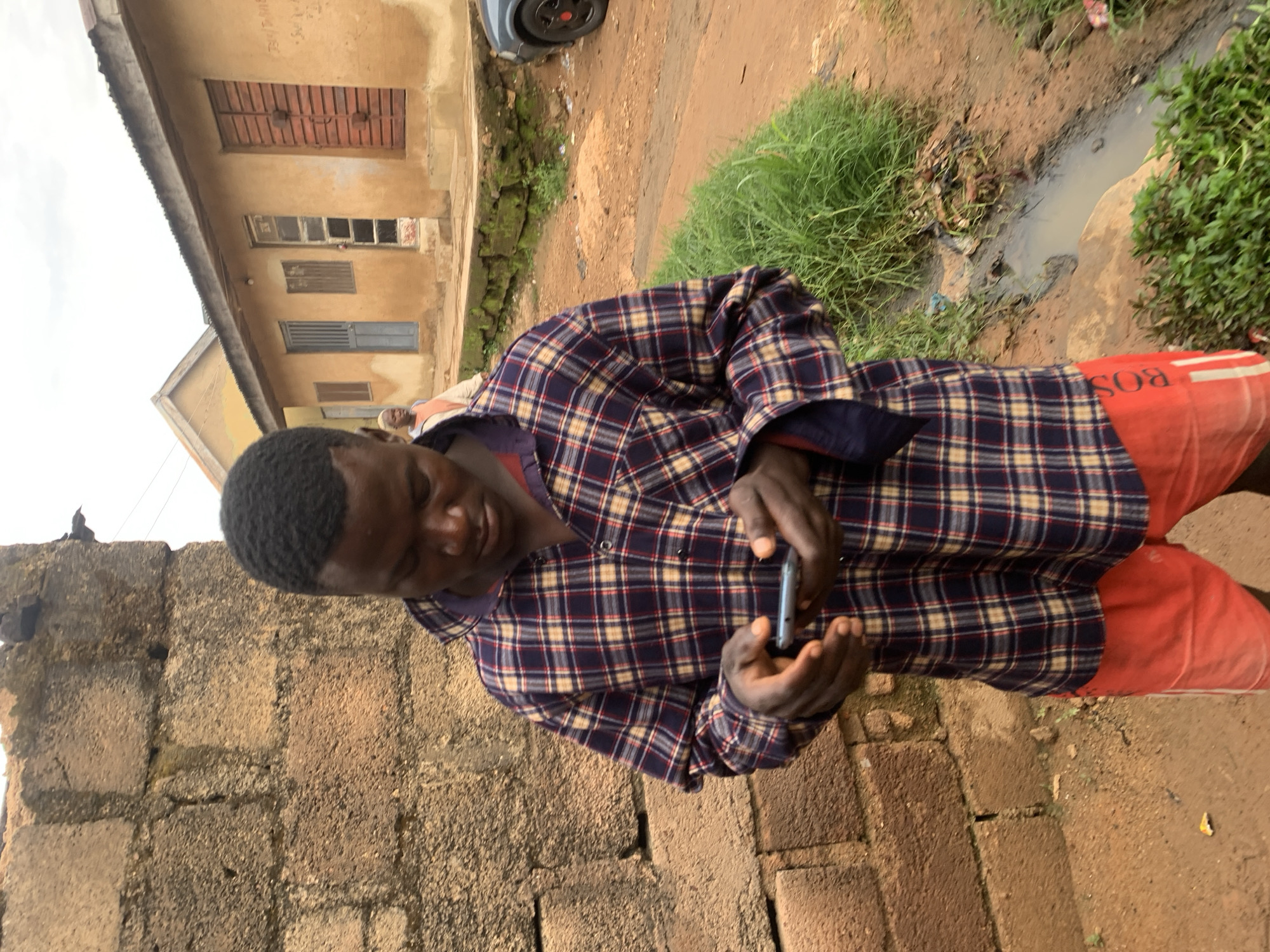 The image depicts an outdoor scene in Jos, Nigeria. In the foreground, a dark-skinned male with short, dark hair is positioned centrally, looking down intently at a silver-colored smartphone held in both hands. He wears a long-sleeved plaid shirt featuring dark blue, red, and beige checks, and red shorts with vertical white stripes and the text "BOSS" on the left leg.
To his left, a rough, weathered wall constructed from grey-brown concrete blocks serves as a partial background. In the middle ground, behind the subject's left shoulder, a light-skinned individual in light clothing is partially visible, walking rightward.
The background primarily consists of two single-story structures with mud-colored walls and corrugated metal roofs. The closest building, directly behind the subject, features multiple access points including a light blue metal door, a brown wooden window, a broken glass door, and a prominent reddish-brown wooden double door. Some faint writing is discernible on its wall.
The ground is predominantly reddish-brown earth, interspersed with patches of green grass and small plants. A shallow, narrow drainage ditch containing stagnant, murky water and debris runs through the middle ground. A dark-colored vehicle is partially visible on the far right, showing a wheel with a red hubcap. The sky is overcast.