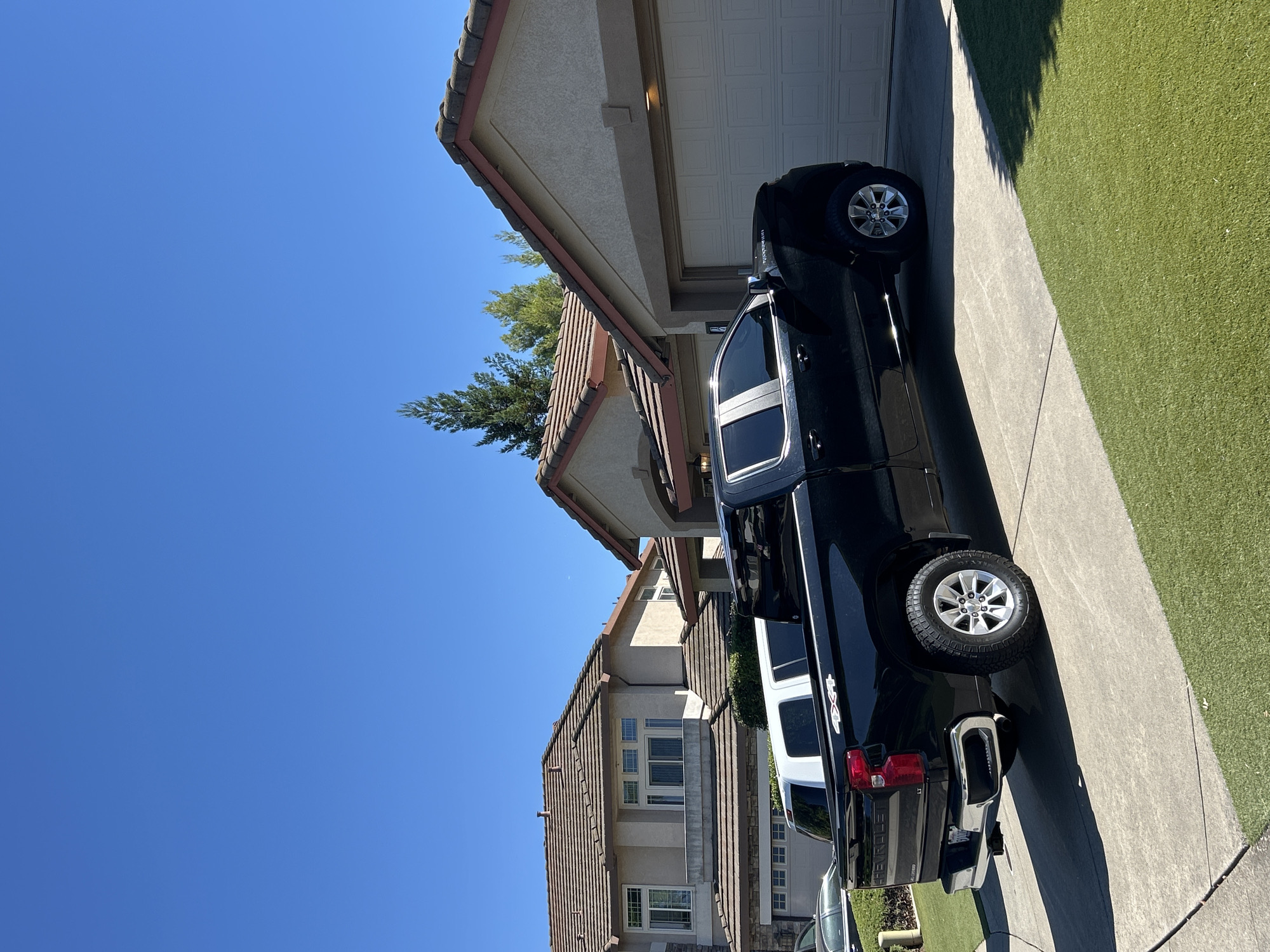 This is a daylight, eye-level, outdoor, medium shot of a black Chevrolet Silverado pickup truck parked in a driveway in front of a residential property. The truck is parked parallel to the house. The truck has a crew cab and a short bed. It has chrome trim around the windows, a chrome bumper, and chrome wheels. The truck bed has "4x4" decals on the sides. A white vehicle is parked behind the Silverado. The driveway is paved with concrete and bordered by artificial turf. In the background, there are two large, tan-colored houses with Spanish tile roofs. The sky is clear and blue. The scene is located in Roseville, United States of America.