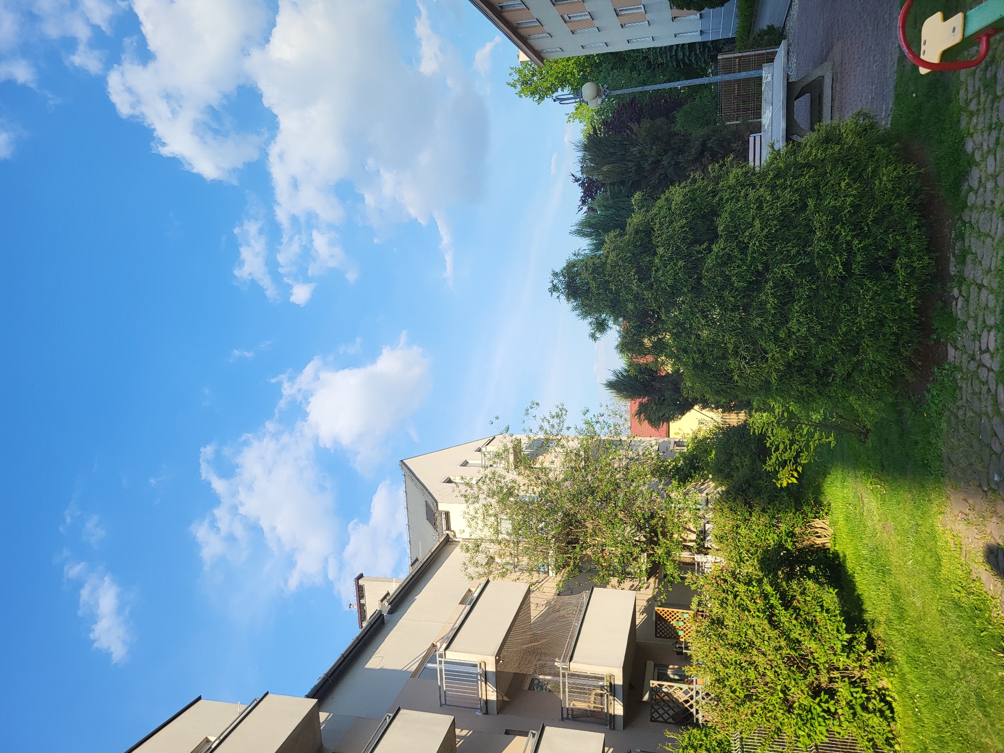 The image presents an outdoor view in an urban setting, likely a residential area. The sky, taking up a significant portion of the frame, is a bright blue with scattered white clouds. Two buildings flank the sides of the image. The building on the bottom left is a multi-story apartment complex with balconies, rendered in light gray tones. The building on the top right is shorter, with a more traditional architectural style. In the foreground and midground, there is abundant greenery including lawns, hedges, and trees. A small section of a playground apparatus is visible in the top right corner. A stone pathway cuts through the greenery.