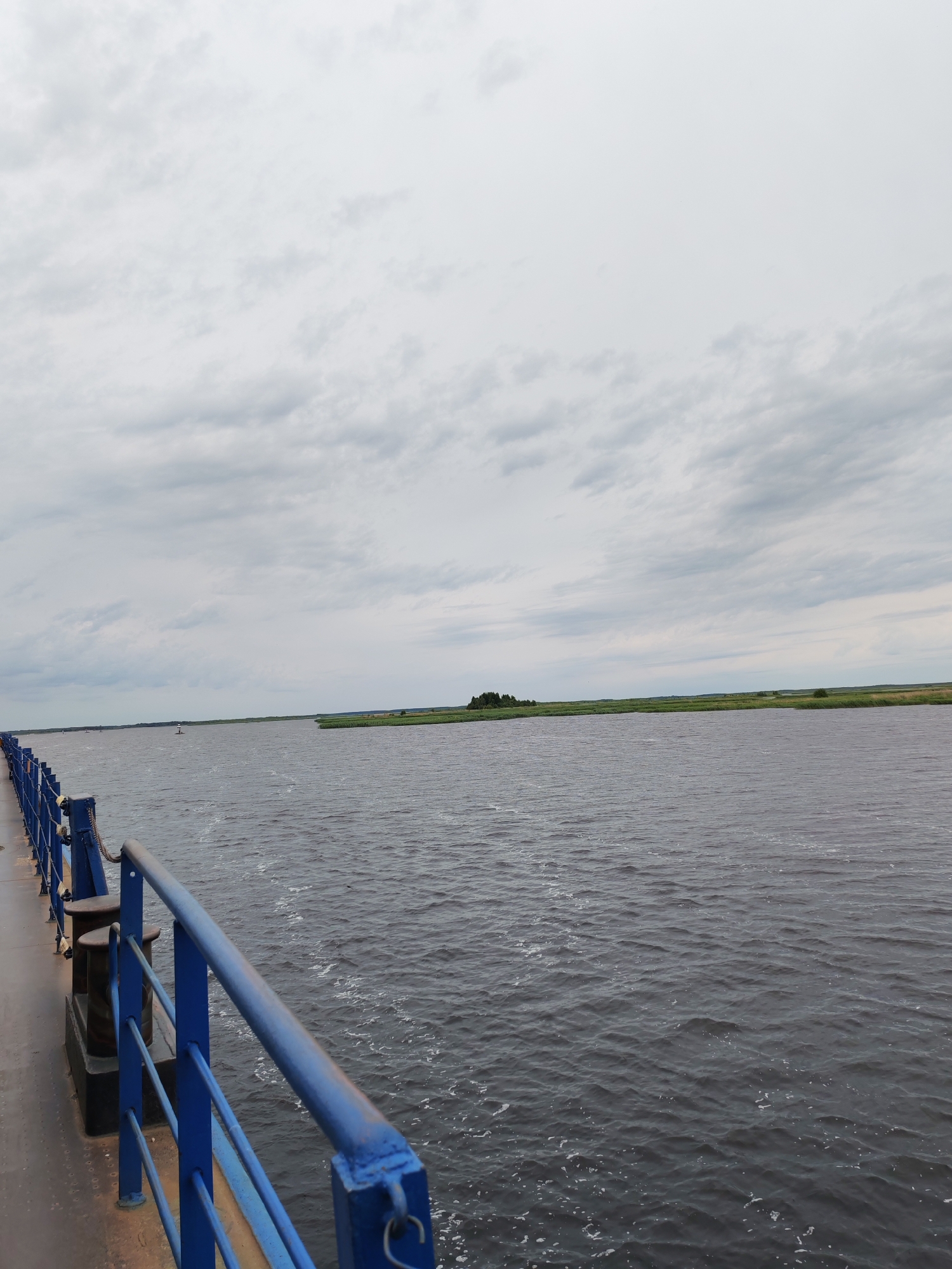 This image depicts a wide, calm body of water, likely a river or a lake, under an overcast sky. In the foreground, a blue metal railing extends along a pier or a walkway. The railing is composed of vertical posts and horizontal bars, with a thick blue top rail. Mooring bollards are visible behind the railing on the walkway, suggesting this is a place where boats might dock. The water's surface is dark and rippled, with small white foam patches, indicating gentle movement.
In the middle distance, the water stretches to a distant shoreline. The shoreline is lined with lush green reeds and vegetation, interspersed with a few trees. A small, dark green island with trees is visible further out in the water on the left side of the frame. On the right side, the shoreline appears flat and extensive, with more reeds and some scattered trees.
The sky is filled with thick, grey clouds, characteristic of an overcast or partly cloudy day. The lighting is diffused, creating a subdued atmosphere. There are no people visible in the image. The scene appears to be a natural landscape viewed from a man-made structure, likely in the daytime, given the amount of light. The location is identified as Bereg, Russia. The overall mood is one of tranquility and natural beauty, with a hint of industrial presence from the pier railing.