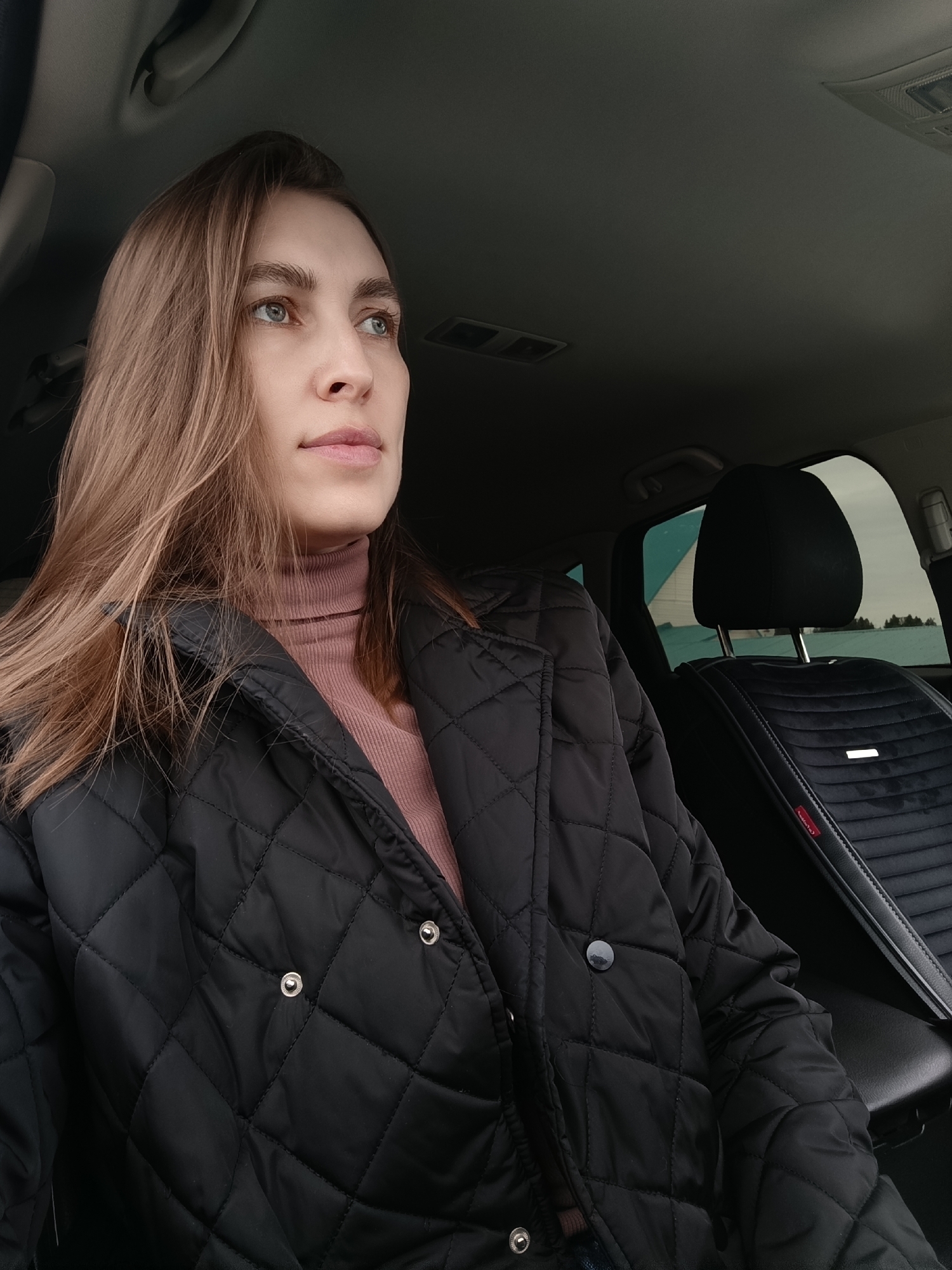 A woman is seated inside a car, wearing a black quilted jacket over a pink turtleneck sweater. She is looking out the window. The car's interior is visible, including the headrest of the driver's seat, which has a black quilted pattern. Through the window, a snowy landscape with trees can be seen. The photo appears to be taken in Фоки, Russia based on the latitude and longitude information.