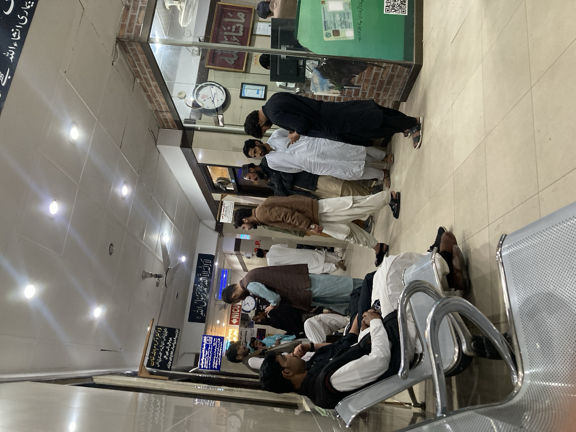 The image shows a waiting area, likely in a hospital or government office in Quetta City Tehsil, Pakistan. A line of men are queuing up at a service window. Some men are seated in the foreground on metal chairs. Several signs in Urdu are posted on the wall, including one that reads "NICU" and another with religious text. The environment appears to be indoors with fluorescent lighting.