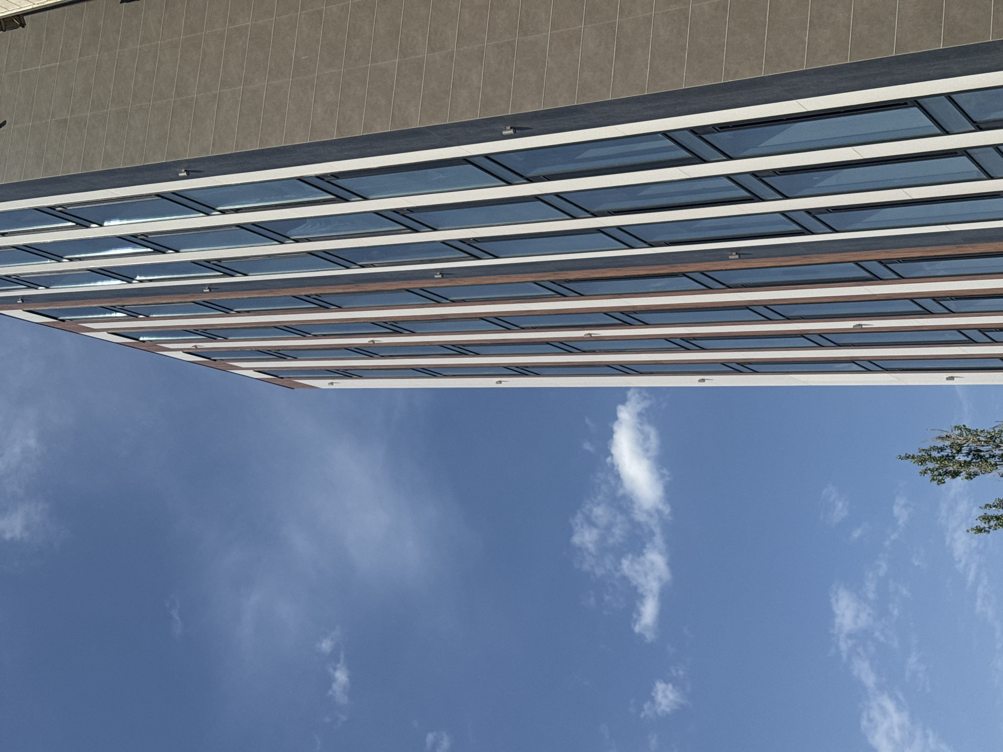 A high-angle exterior shot shows the side of a modern, multi-story building with a facade of alternating dark grey, white, and wood-toned vertical panels. The building has a repetitive pattern of tall, narrow windows with dark frames. The sky is a clear, vibrant blue with scattered wispy white clouds. The lower left corner of the image shows the tops of green trees. The scene appears to be an urban environment, possibly in Bishkek City, Kyrgyzstan, given the architecture and vegetation. There are no people visible.