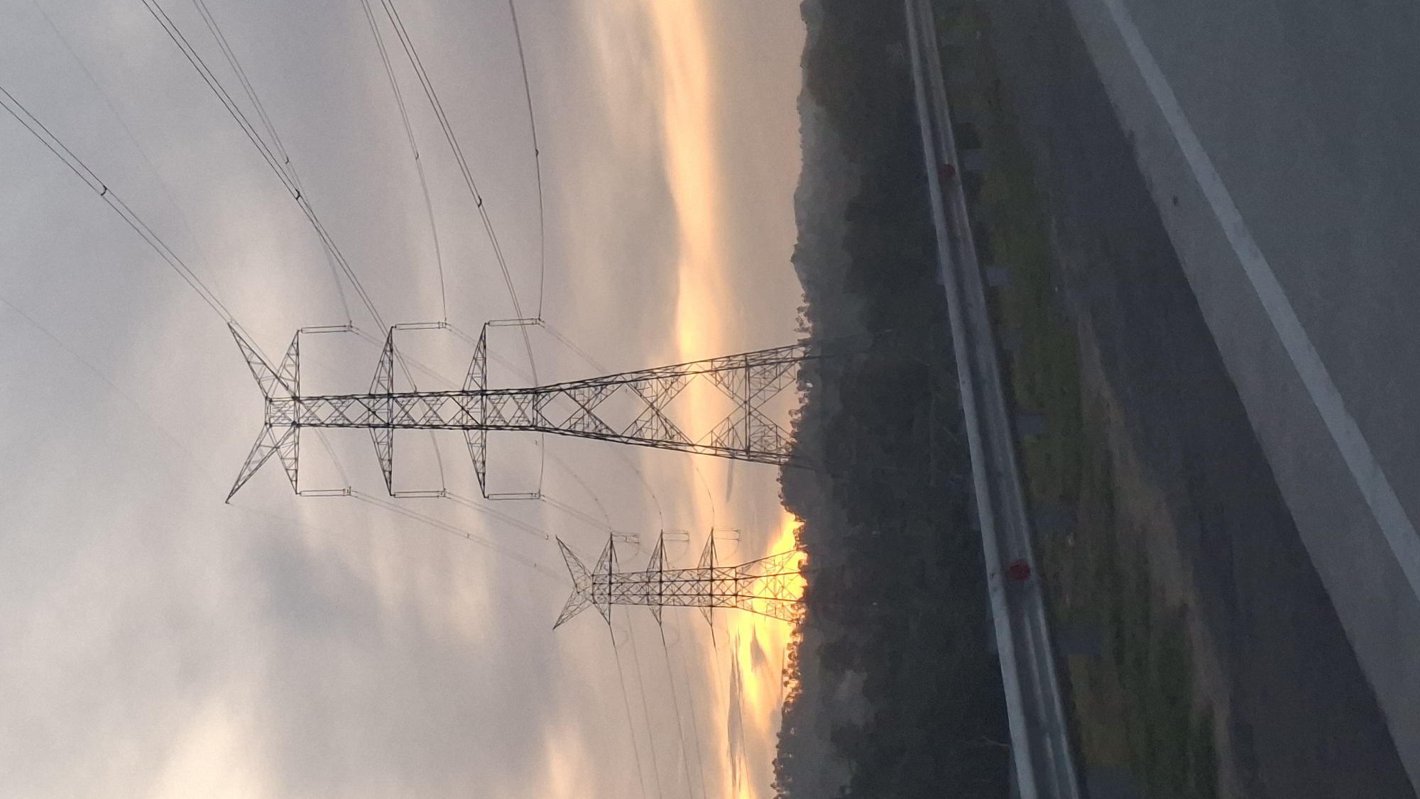 Two tall, lattice-style electrical transmission towers dominate the frame, silhouetted against a cloudy sky with a warm, orange glow of sunrise or sunset peeking through the clouds between the towers and behind a line of dark green trees and hills. The towers are a dark gray, almost black, color. The left tower is slightly smaller and set further back than the one on the right, which is more prominently in the foreground. Multiple power lines extend from the tops of the towers and stretch across the sky. In the foreground, a gray metal guardrail runs horizontally across the lower part of the image, separating the road from the grassy embankment behind it. The road surface is dark asphalt with a white line marking its edge. The scene appears to be set in a rural or semi-rural area, possibly on the outskirts of the city of Lamtamot, Indonesia, given the presence of the electrical infrastructure and the natural landscape. There are no people visible in the image.