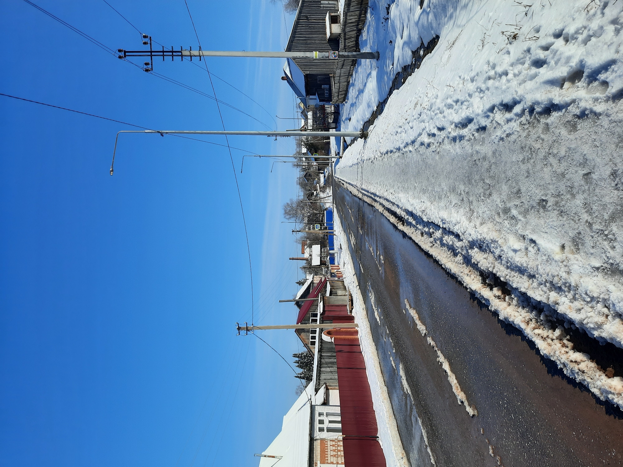 The image depicts a street scene in the village of Krasnogorskoye, Russia, during winter. The street is partially covered with melting snow and ice. The road surface appears wet. Along the sides of the street, there are snowbanks with dark brown sediment visible at the base. The sky is clear and blue.
On either side of the street, there are residential buildings of varying heights and architectural styles, mainly constructed from wood. Some buildings have red, brown, or gray fences. Power lines and street lamps run along the street. In the distance, leafless trees are visible. No people are present.