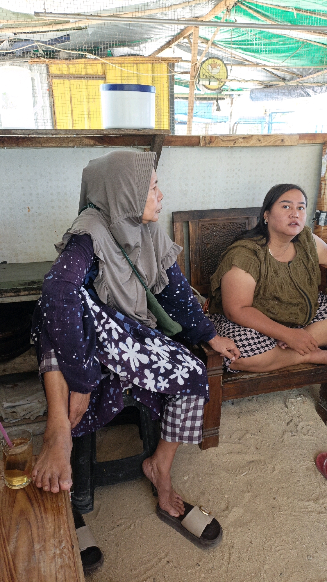 Two women are seated on wooden chairs on a sandy surface under a makeshift roof structure. The woman on the left, wearing a gray hijab and a dark purple dress with white floral patterns and checkered pants, has her left leg extended forward. A glass of iced tea sits on a wooden table in front of her. The woman on the right is wearing a green top and a black and white patterned skirt. She is looking towards the left. The background reveals a white wall and a yellow structure behind a metal grid. The overall scene suggests a casual outdoor setting in a location like Cikal, Indonesia.