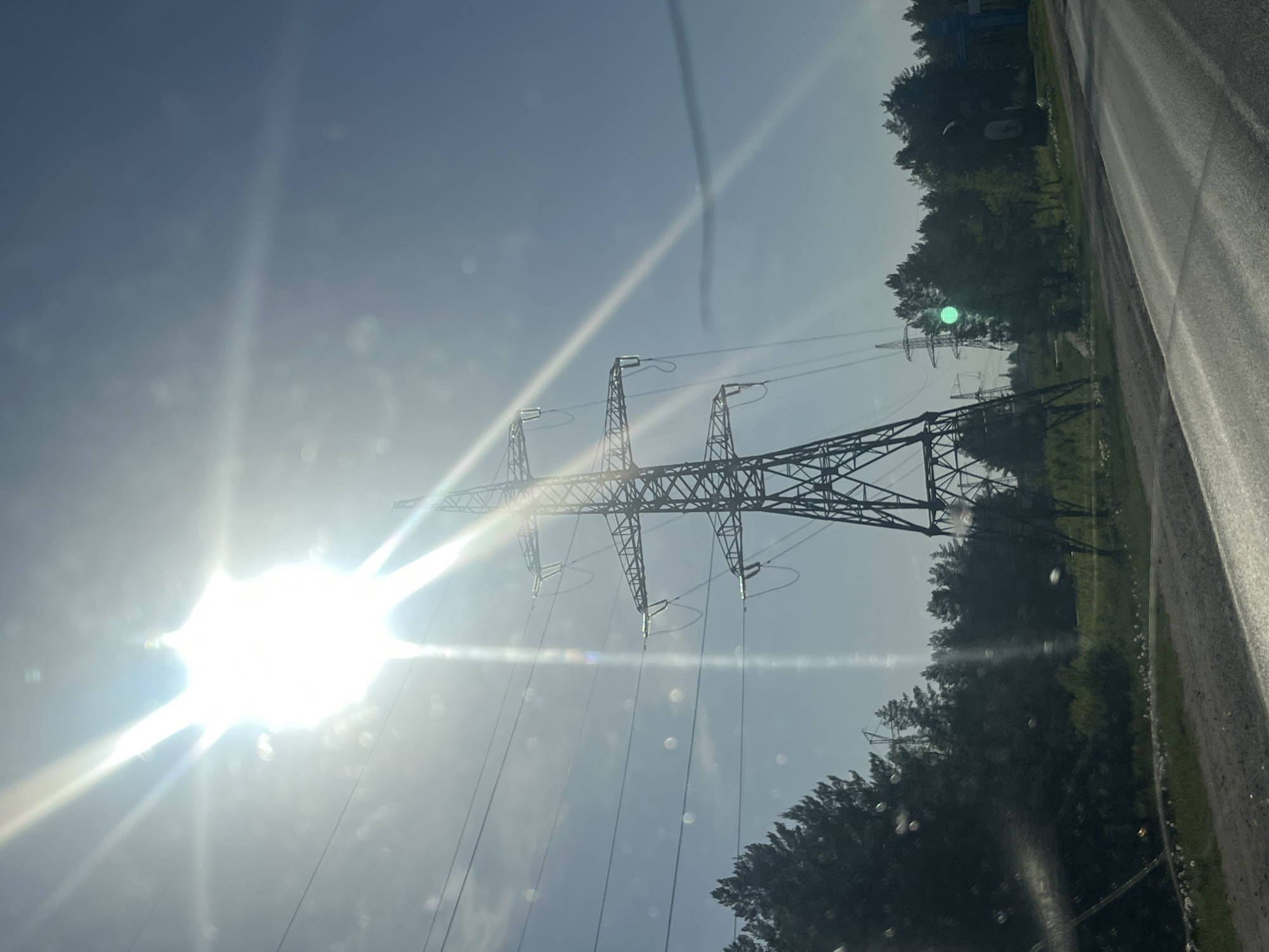 The image depicts a bright, sunlit scene with power lines and pylons in the foreground. The pylons are of a lattice construction, suspending several transmission lines. Dense trees line the right side of the frame, bordering what appears to be a road. The sun is located on the left side of the frame, obscuring details with its glare. The location is Noviy, Russia. No people, actions, or interactions are visible.