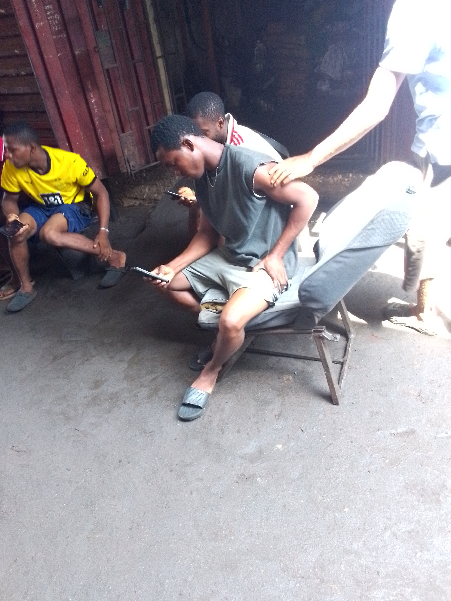 An outdoor scene captures four young men in what appears to be an urban setting in Onitsha, Nigeria. Three of the men are prominently visible, seated on various informal chairs and stools, each engrossed in their mobile phones.
In the foreground, a man wearing a grey t-shirt, grey shorts, and dark slide sandals sits on a simple metal chair, focused intently on his smartphone. To his left, another man in a bright yellow football jersey with "BVB 1&1" text and blue shorts is also seated, holding a phone. His left leg is crossed over his right, and he wears dark sandals. Behind him, another man's head and shoulders are visible, also looking down, presumably at a phone. A fourth person's arm and lower body are visible on the far right, also holding a phone and standing or sitting on a higher surface, with a hand resting on the shoulder of the man in the grey t-shirt.
The setting features a large, dark red corrugated metal structure in the background, possibly part of a building or a shipping container, with some indistinct writing visible on its surface. The ground is rough, grey concrete or pavement. The lighting is uneven, with some areas brightly lit by what appears to be sunlight, while others, particularly in the background, are in deep shadow. The overall impression is one of casual social gathering in a relaxed, informal environment during the daytime, with the primary activity being engagement with personal electronic devices.