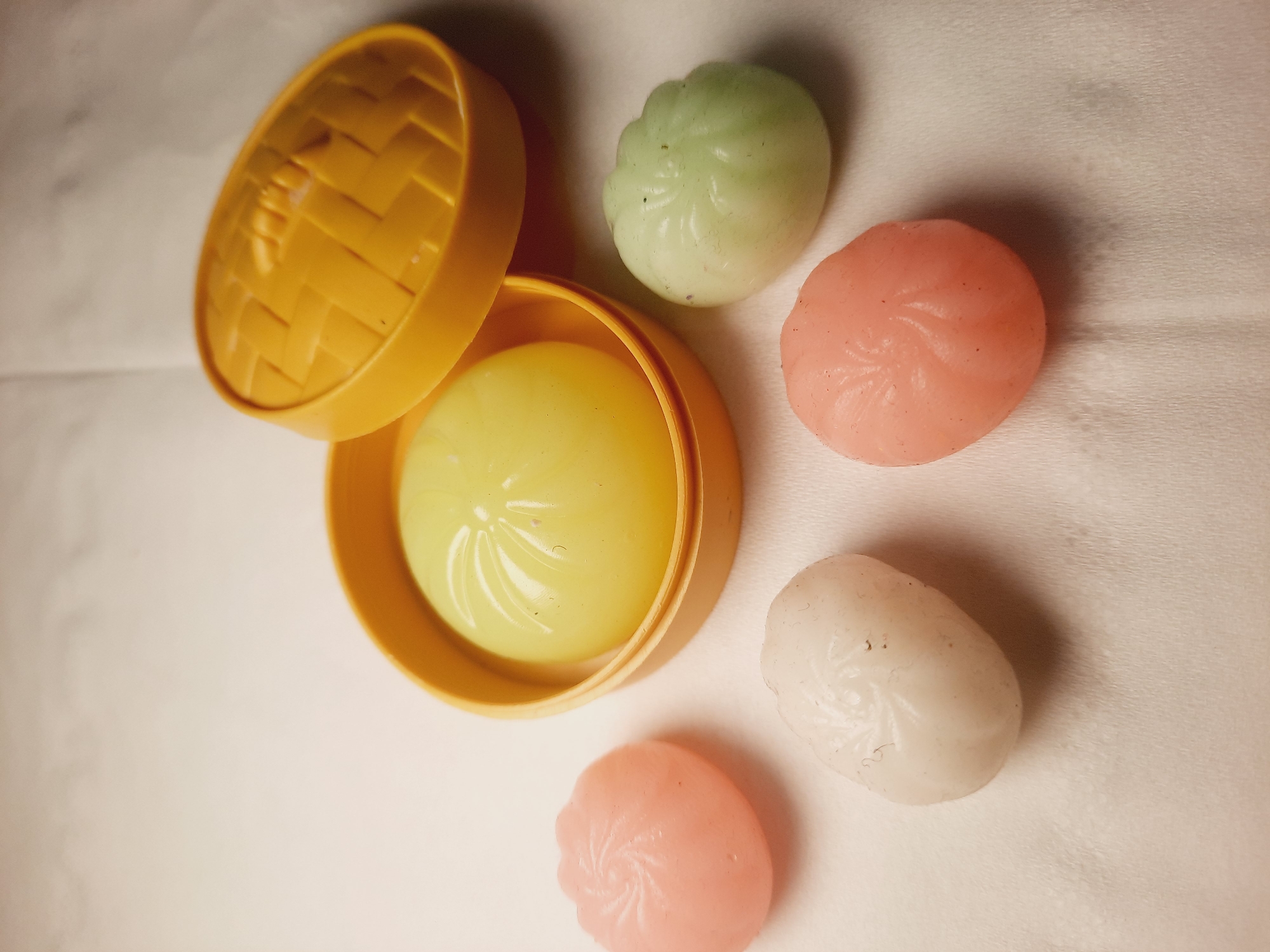 Five small, stylized, bun-shaped objects are arranged on a textured white surface. One yellow object, with a woven basket-like texture on its lid, is open, revealing a smooth, pale yellow, bun-shaped item inside. To the left of this container is a pink bun-shaped object. Below this and to the left is a white bun-shaped object. To the right of the white object is another pink bun-shaped object. To the right of the yellow container is a pale green bun-shaped object. The objects appear to be toys or decorative items, possibly resembling steamed buns. The lighting suggests an indoor setting with a soft overhead light source. There is no indication of the city of North Jakarta, Indonesia in the image.
