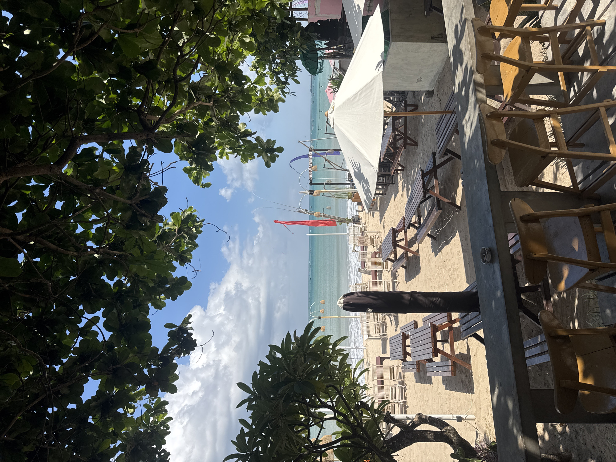 This is a daytime, eye-level, medium shot of a beachside seating area with the ocean in the background. The scene is located in Kuta, Indonesia. In the foreground, a concrete counter with several wooden bar stools is visible. Behind the counter, picnic tables and benches are arranged on the sand. Further back, rows of lounge chairs are set up, facing the ocean. The water is calm and turquoise, with a clear sky and scattered clouds. Lush green trees frame the top of the image, casting shadows on the scene. There are no people actively engaged in the scene, but the presence of tables and chairs suggests a public space, likely a cafe or restaurant. The overall atmosphere is relaxed and inviting, with a focus on outdoor leisure.