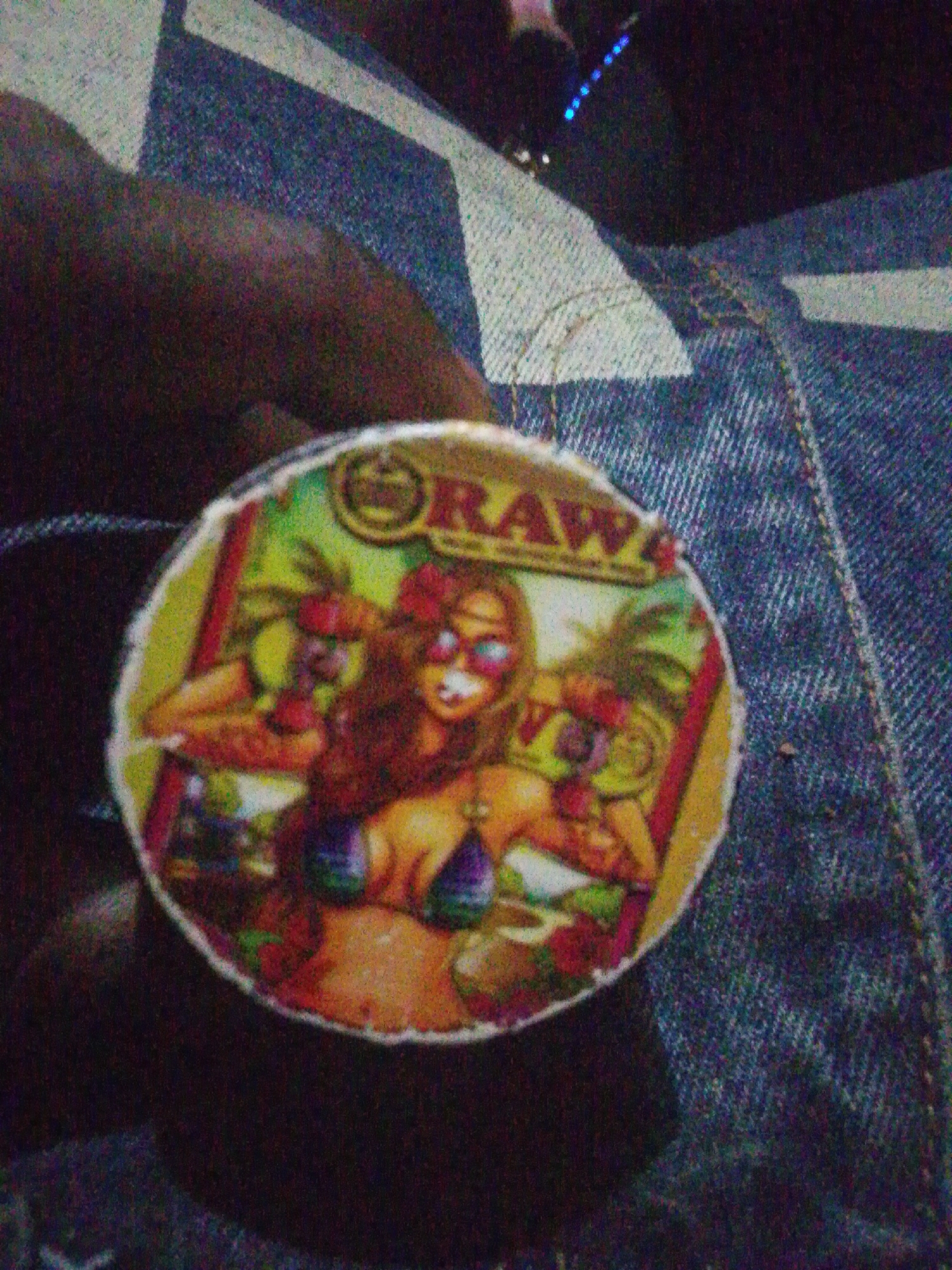 A forensic examination of the image reveals a circular object, approximately 7-8 cm in diameter, held by a person's hand. The object features a vibrant, cartoonish illustration with a tropical theme. At the top, the word "ORAWA" is visible in bold, stylized lettering, with a small crown or emblem positioned above the letter 'O'. The central figure is an anthropomorphic female character with brown hair, wearing red sunglasses and a bikini top in shades of purple and green. She has a red flower in her hair. Flanking her on either side are two smaller, stylized figures resembling individuals with green, spiky "palm tree" hair, also wearing sunglasses. The background of the illustration includes palm trees and what appear to be food items, possibly burgers or sandwiches, suggesting a beach or vacation setting. The color palette of the illustration is bright, featuring greens, yellows, reds, and blues.
The circular object is grasped from the lower right by a dark-skinned hand, with fingers partially visible. The immediate background consists of blue denim fabric, exhibiting a distinctive weave pattern and visible stitching, consistent with a pair of jeans. In the upper right portion of the image, faint blue lights or reflections are discernible against a dark background, but are too indistinct to identify further. The overall image quality is characterized by significant digital noise and blur, suggesting low-light conditions and/or a low-resolution camera.
The location is undetermined and cannot be ascertained from the visual information provided within the city of Unknown, Unknown.