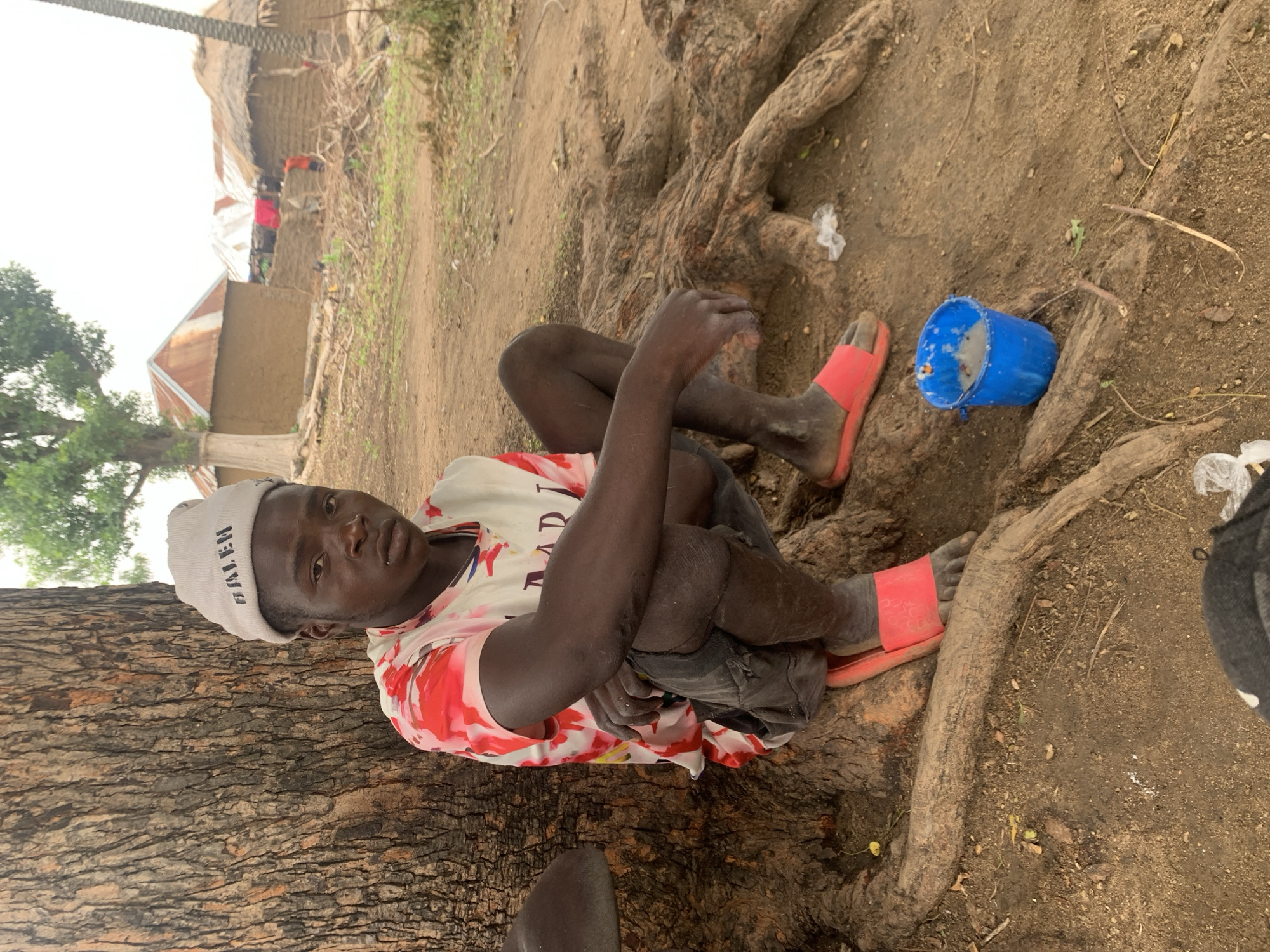 A young Black male, appearing to be in his late teens or early twenties, is crouched next to the base of a large tree, in what appears to be the city of Dull A, Nigeria. He is wearing a white beanie with the word "BALEN" printed on it in black lettering, a white t-shirt with red and pink abstract patterns, and dark shorts. He has on red slide sandals. His knees are up and his hands are resting on his knees. To his right, a small blue bucket contains a light-colored liquid or substance. The ground is dirt and is covered with tree roots. In the background, several rustic buildings with thatched or corrugated metal roofs are visible, suggesting a rural or semi-rural setting within Dull A, Nigeria. The sky is overcast.