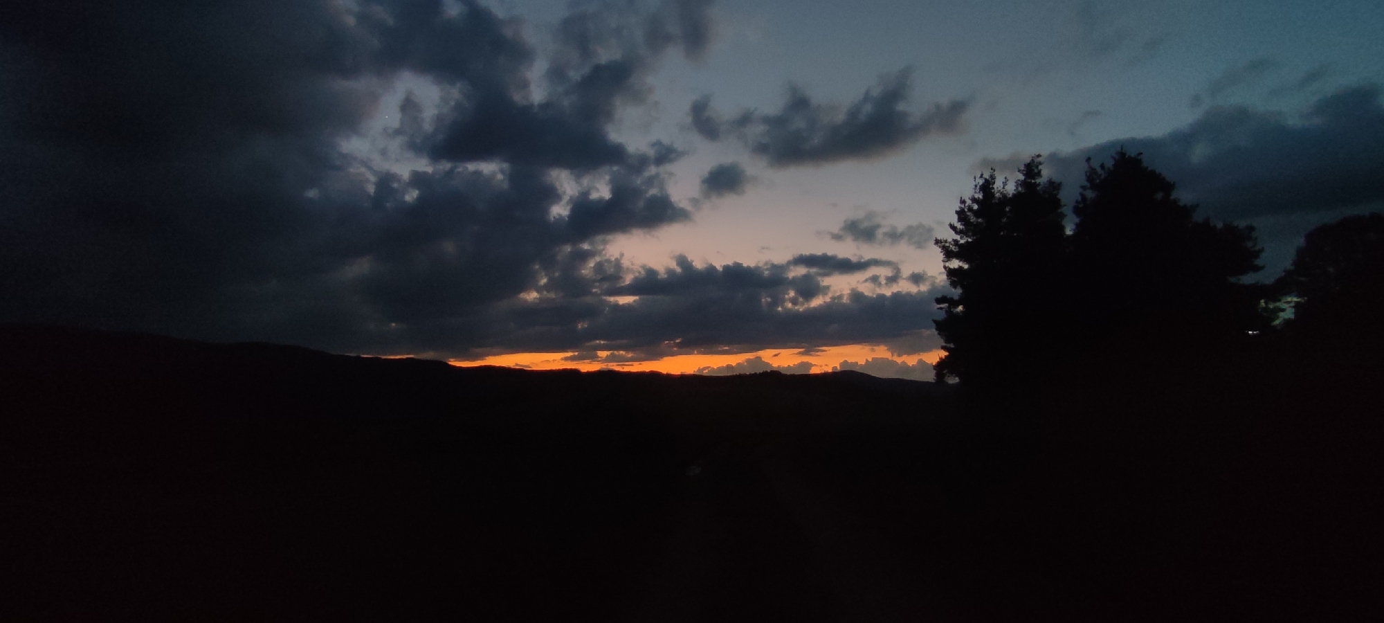 The image depicts a twilight landscape scene in Gutsal, Bulgaria, characterized by a distinct sunset or dusk. The upper portion of the frame is dominated by a heavy, dark cloud cover, particularly dense on the left. A horizontal band of intense orange light, indicative of a low sun, is visible across the mid-ground, separating the shadowed terrain from the upper sky. Above this orange band, the sky transitions to various shades of dark grey and desaturated blue, with a small, bright white punctiform object, consistent with a celestial body, faintly visible within the lighter cloud area in the upper right quadrant. The lower half of the image presents a dark silhouette of rolling hills or mountains along the horizon, situated directly beneath the orange light. The immediate foreground and lower mid-ground are rendered in deep shadow, appearing as an undifferentiated dark mass of terrain and vegetation. On the far right edge, a prominent, dark silhouette of one or more coniferous trees, likely pines, extends vertically, partially obscuring the sunset. No human figures, animals, man-made objects (beyond potential distant terrain features), actions, or interactions are discernible.