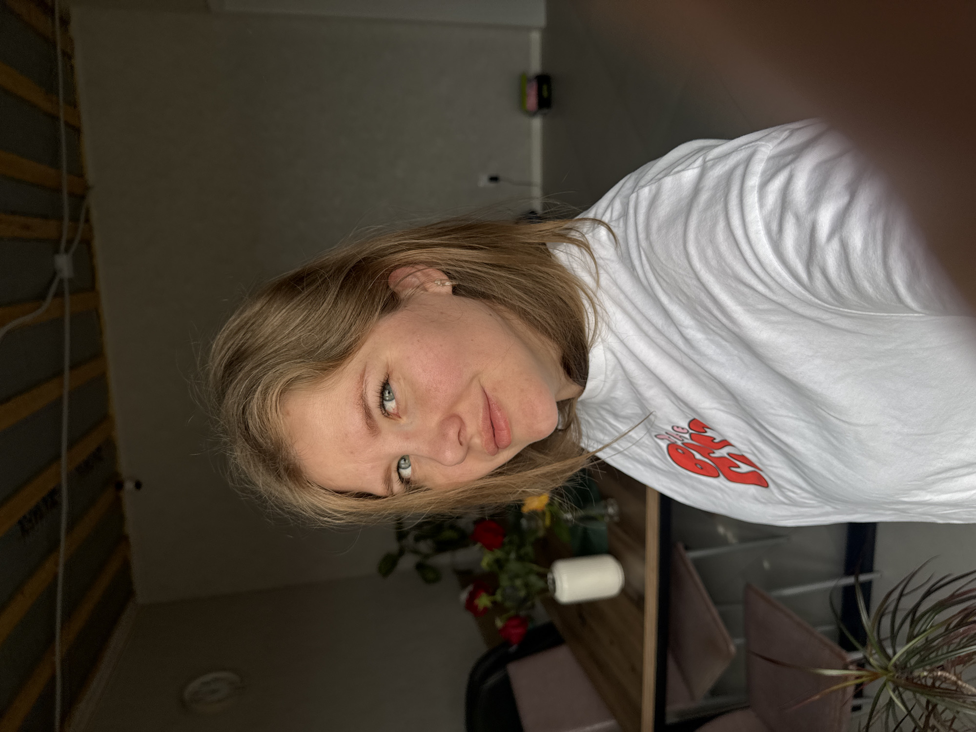 The image is a close-up selfie of a fair-skinned young woman with shoulder-length blonde hair and light eyes. She is wearing a white t-shirt with a red design. In the background, there is a shelf with a roll of paper towels and a vase of red and yellow flowers. Additionally, there are a few houseplants. The location is a dimly lit interior room in Gornyi (Dvurechenskoe S/P), Russia.