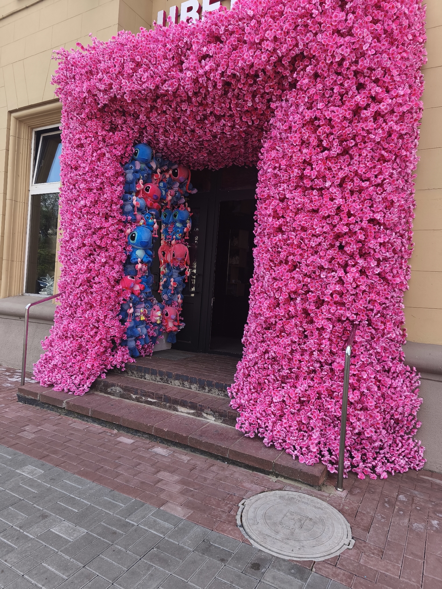 The image depicts an entryway to a building, heavily decorated with artificial pink flowers. The flowers form an archway over the entrance and cover the facade on either side, cascading down to the ground. Visible within the archway, hanging from what appears to be a display or string, are numerous plush toys, predominantly blue and pink characters that resemble Stitch from Disney's "Lilo & Stitch." The entrance itself is a dark doorway with glass panes and a railing on the left side. Steps lead up to the doorway, made of red bricks. The ground in front of the steps is paved with red and grey bricks laid in different patterns. A manhole cover is visible on the paving stones to the right of the steps. The building facade above the flowers is a light beige color, and a portion of a white sign with the letters "UBP" is visible at the top. The scene appears to be a public area in a city, with the context provided indicating it is located in Minsk, Belarus. There are no people visible in the image.