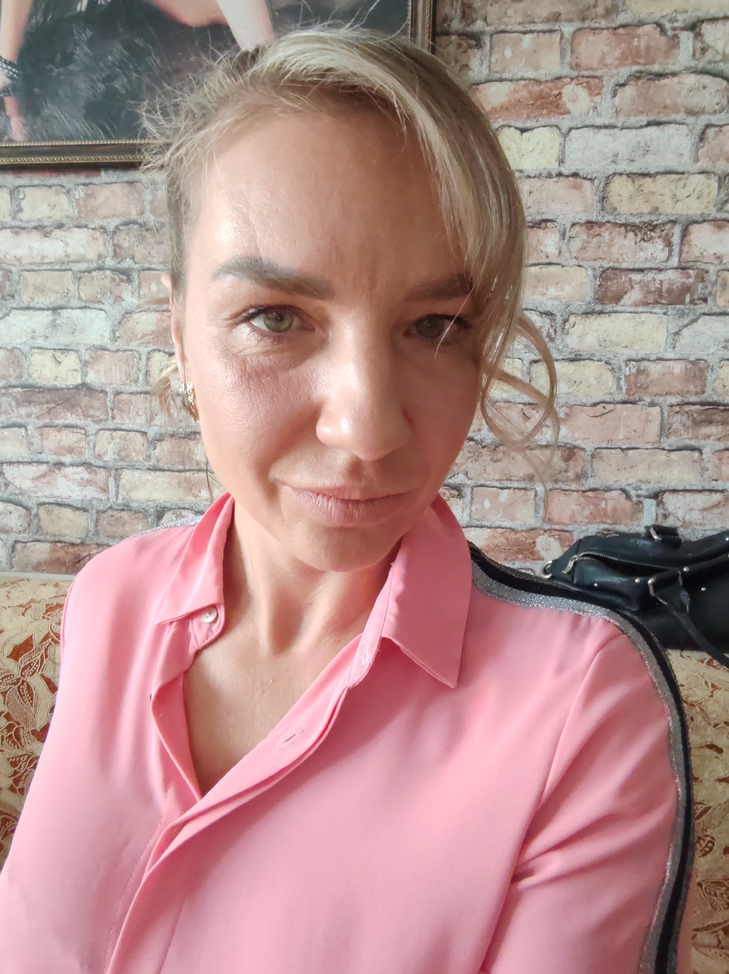 This is a head-and-shoulders selfie of a woman with blonde hair, taken indoors in what appears to be a living room. She is wearing a bright pink collared shirt with silver detailing on the shoulders. Her expression is neutral, with a slight smile. The background consists of a brick wall, a framed picture, and a piece of furniture with a patterned upholstery. A black handbag is visible behind her right shoulder. The lighting suggests it is daytime. The image is a close-up, focusing on the woman's face and upper torso. There is no visible text.