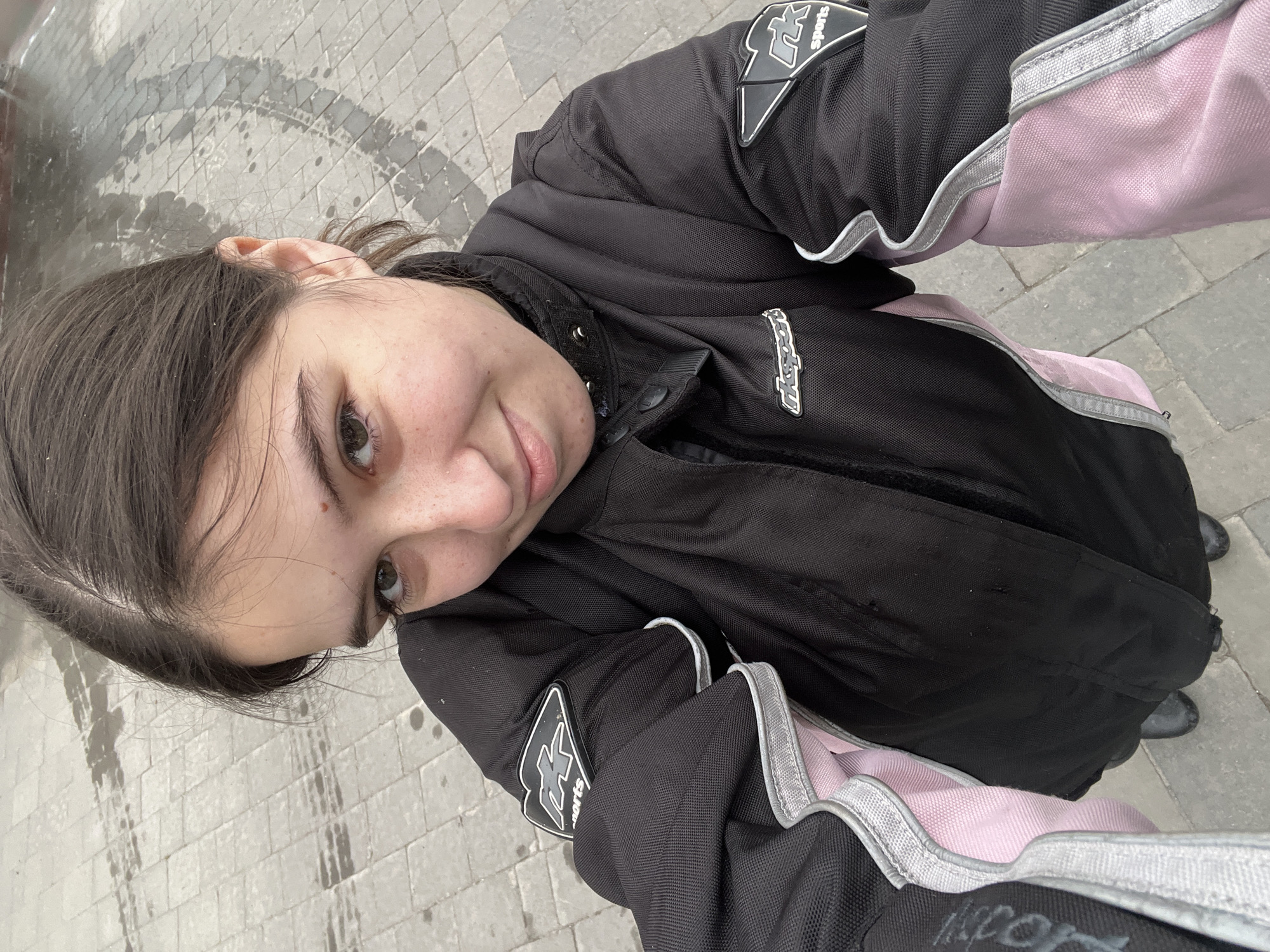 A person wearing a black and pink motorcycle jacket is taking a selfie outdoors. The person is standing on a gray brick sidewalk. The location is possibly Horodok, Ukraine.