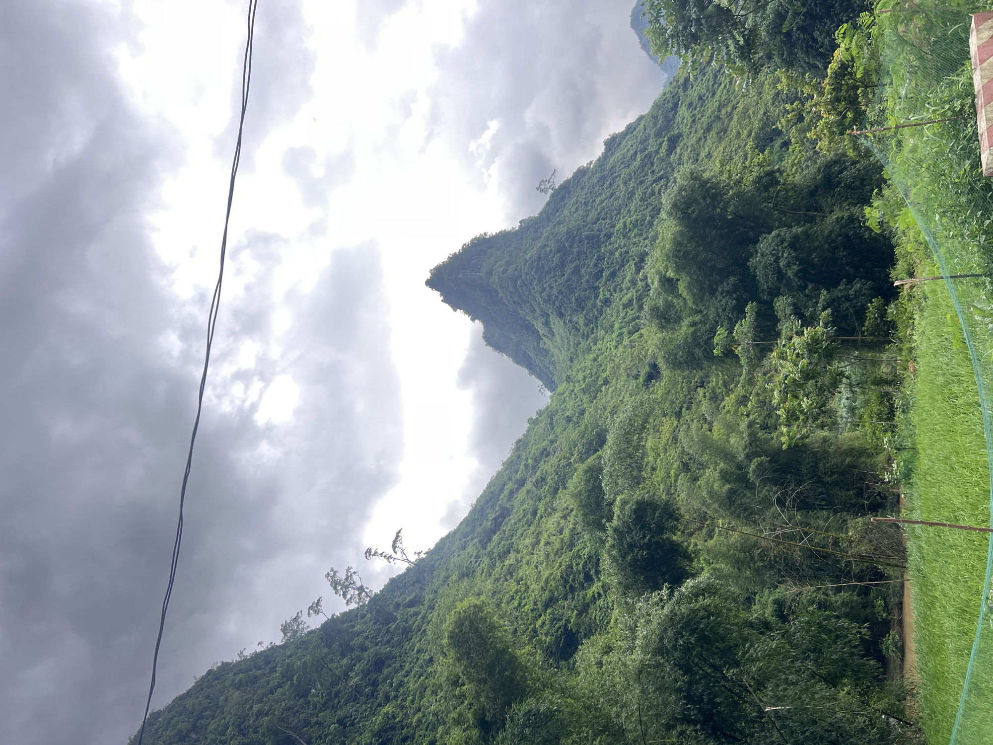A majestic, cloud-covered sky looms over lush, verdant mountains in Cán Tỷ Commune, Vietnam. The mountains are densely covered with trees, with the left side of the image dominated by a steep, tree-covered slope. To the right, a distinct, pointed mountain peak rises sharply. Two dark power lines stretch horizontally across the upper portion of the frame, bisecting the sky. In the foreground, a vibrant green field of grass stretches across the bottom of the image, bordered by a thin, green mesh fence in places. A cluster of bamboo trees can be seen to the left of the foreground field, with a muddy river flowing behind them. The scene is natural and mountainous, with signs of human infrastructure in the form of the power lines and the fence.