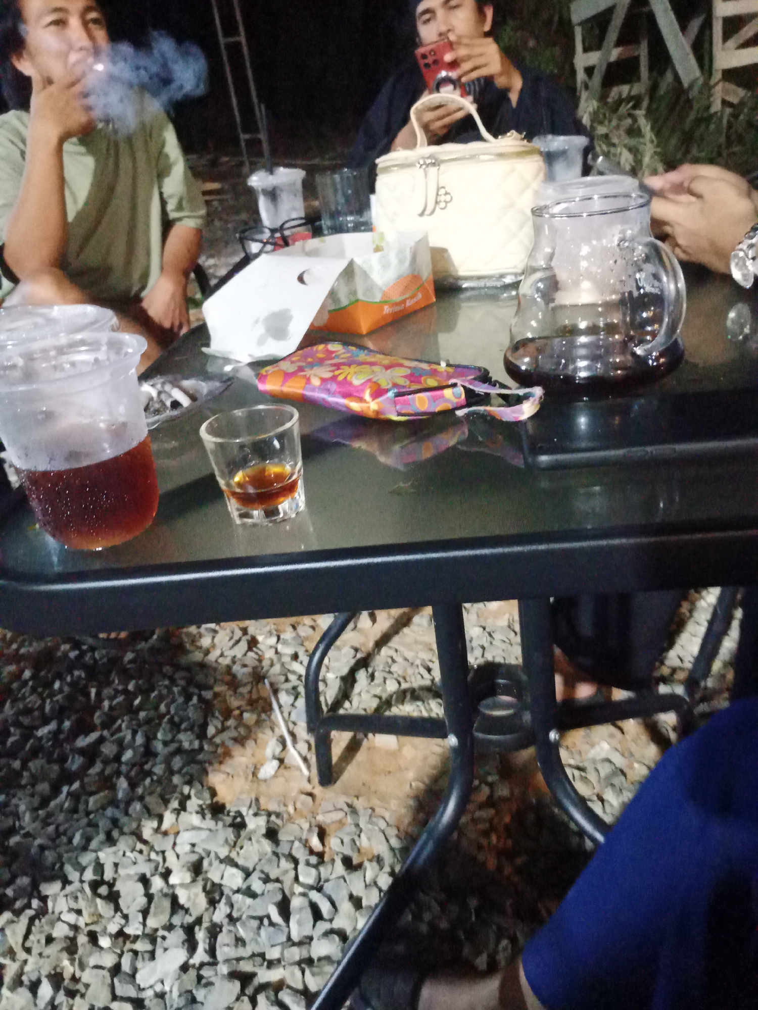 In an outdoor setting at night, possibly a cafe or gathering space in Mekar Jaya, Indonesia, a group of individuals are seated around a glass-topped table. The table is laden with various items including a clear plastic cup filled with a dark beverage, a small shot glass containing a liquid, a clear glass pitcher with dark liquid and steam rising, a quilted clutch bag, and what appears to be a package of snacks. One person is visible in the left of the frame, wearing a light green shirt and smoking, with smoke billowing around their face. Another person is in the background, holding a red mobile phone. The ground surrounding the table is covered in gravel and dirt. The overall atmosphere suggests a casual evening gathering.