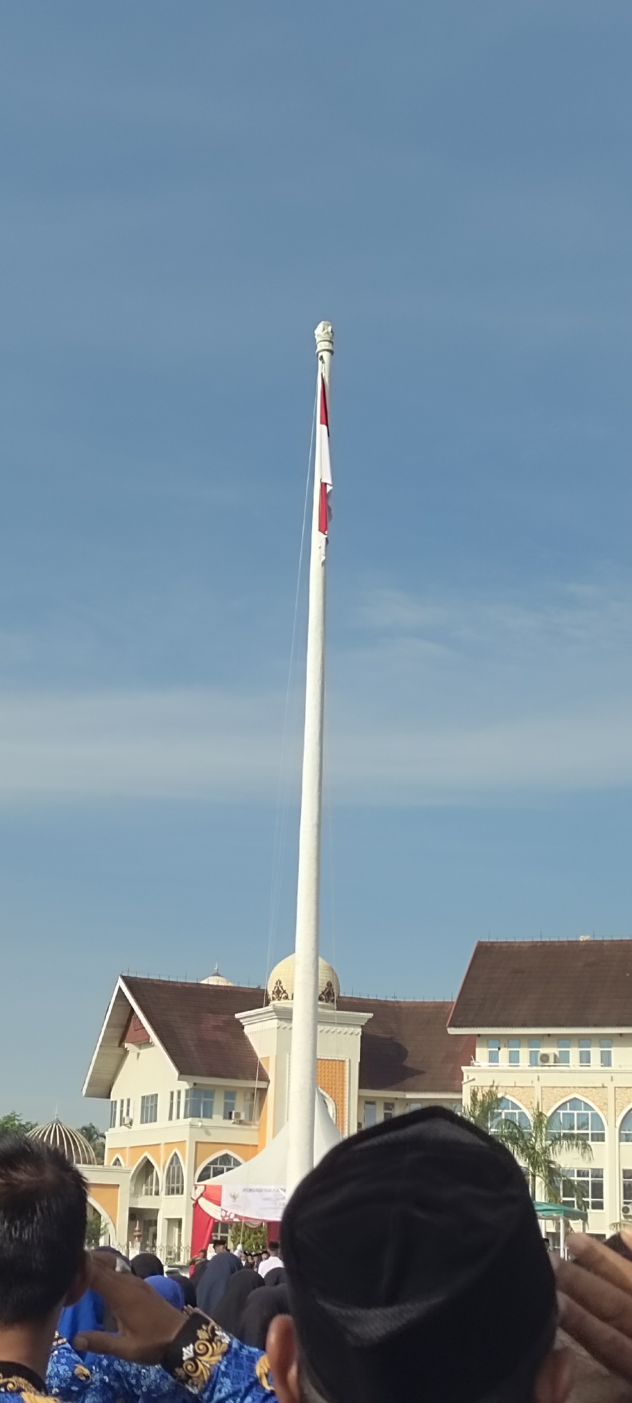 The image depicts a gathering of people in Meunasahaluedrienlhoksukon Barat, Indonesia, likely for a formal event. In the foreground, people are visible, some wearing black caps and blue patterned shirts. They appear to be looking towards a flagpole. In the background, a large building with a brown roof and yellow detailing is visible. A white flagpole stands tall, with a small Indonesian flag waving at the top. The sky is blue with some light clouds.