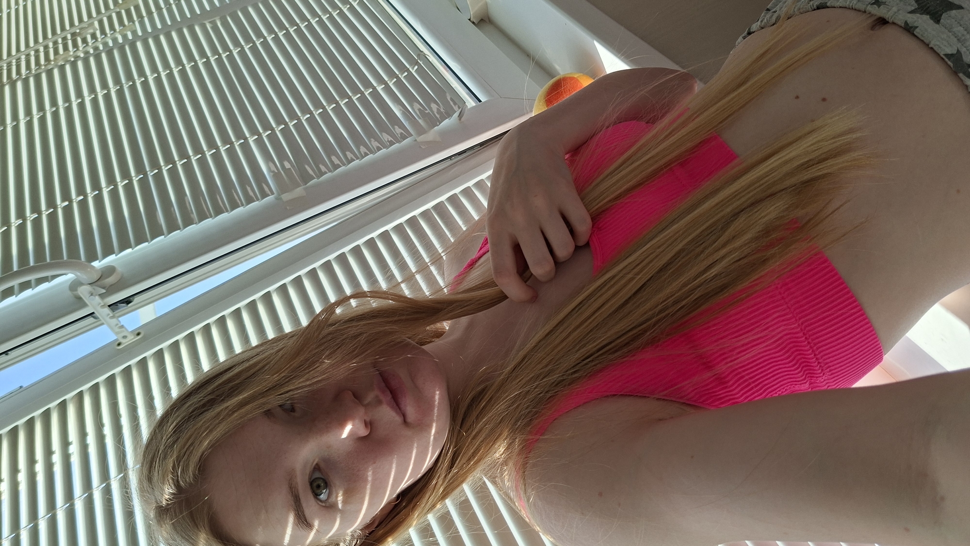 A woman is pictured indoors, seemingly in Chaikovsky, Russia, based on the architectural style visible through the window blinds. She wears a bright pink top and patterned shorts. She is holding her long, blonde hair. The environment appears to be a bright, sunlit room with white window blinds.