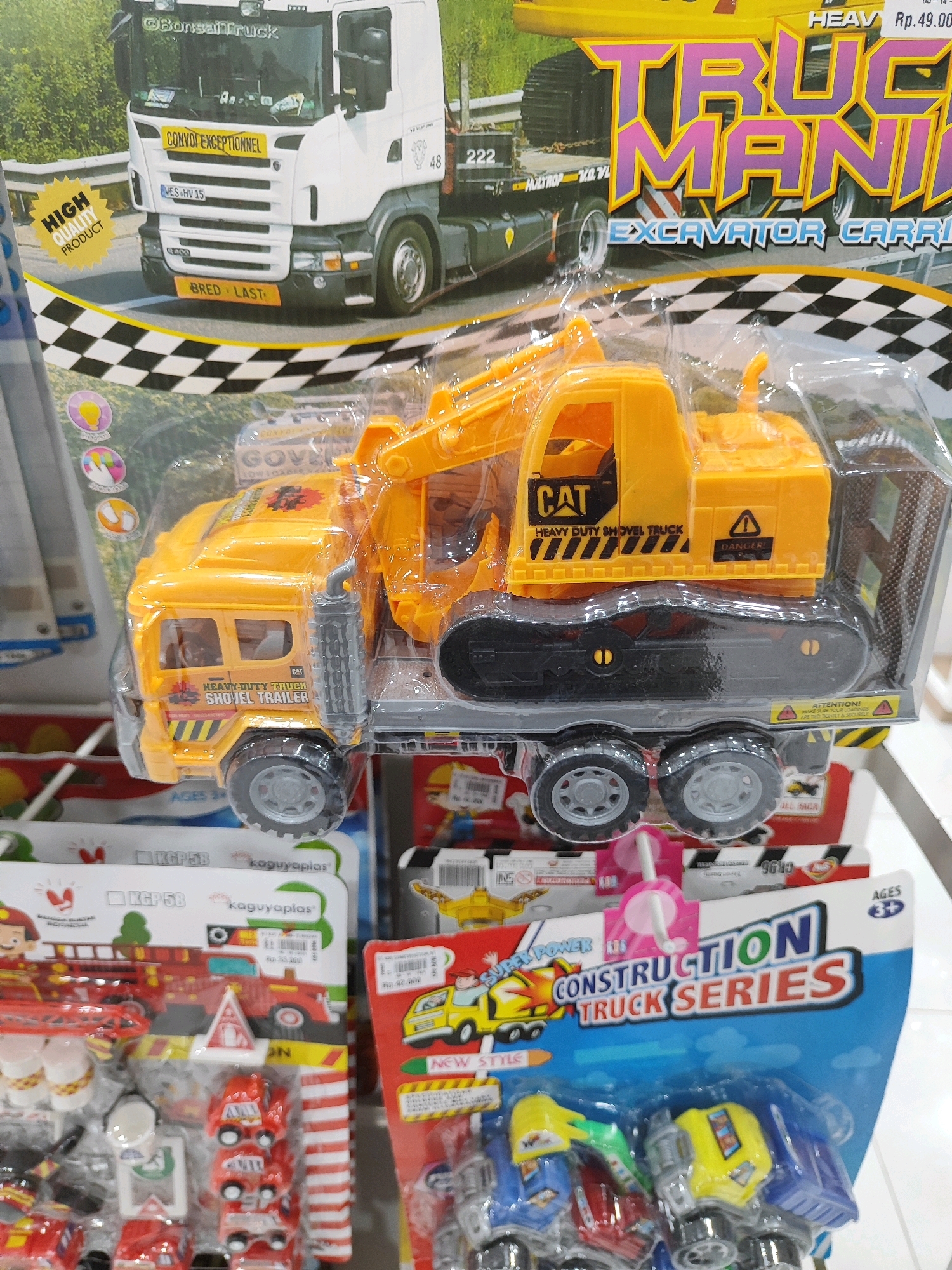 This image captures a retail display of toy trucks, likely in a store. The primary focus is on a large, transparent plastic package containing a yellow and black toy excavator truck with a trailer. The packaging has bold graphics, including the words "TRUCK MANIA" and "EXCAVATOR CARRIER". Next to this, other toy vehicles are visible, including a red fire truck and other smaller construction vehicles. The background shows shelves with more toy packaging. One specific detail in the image is a price tag that reads "Rp. 49.000", indicating Indonesian Rupiah. The question is asking to locate this scene in Sukowiryo, Indonesia, which is not deducible from the image alone. The image displays a commercial environment, suggesting a toy store or a market stall selling toys.