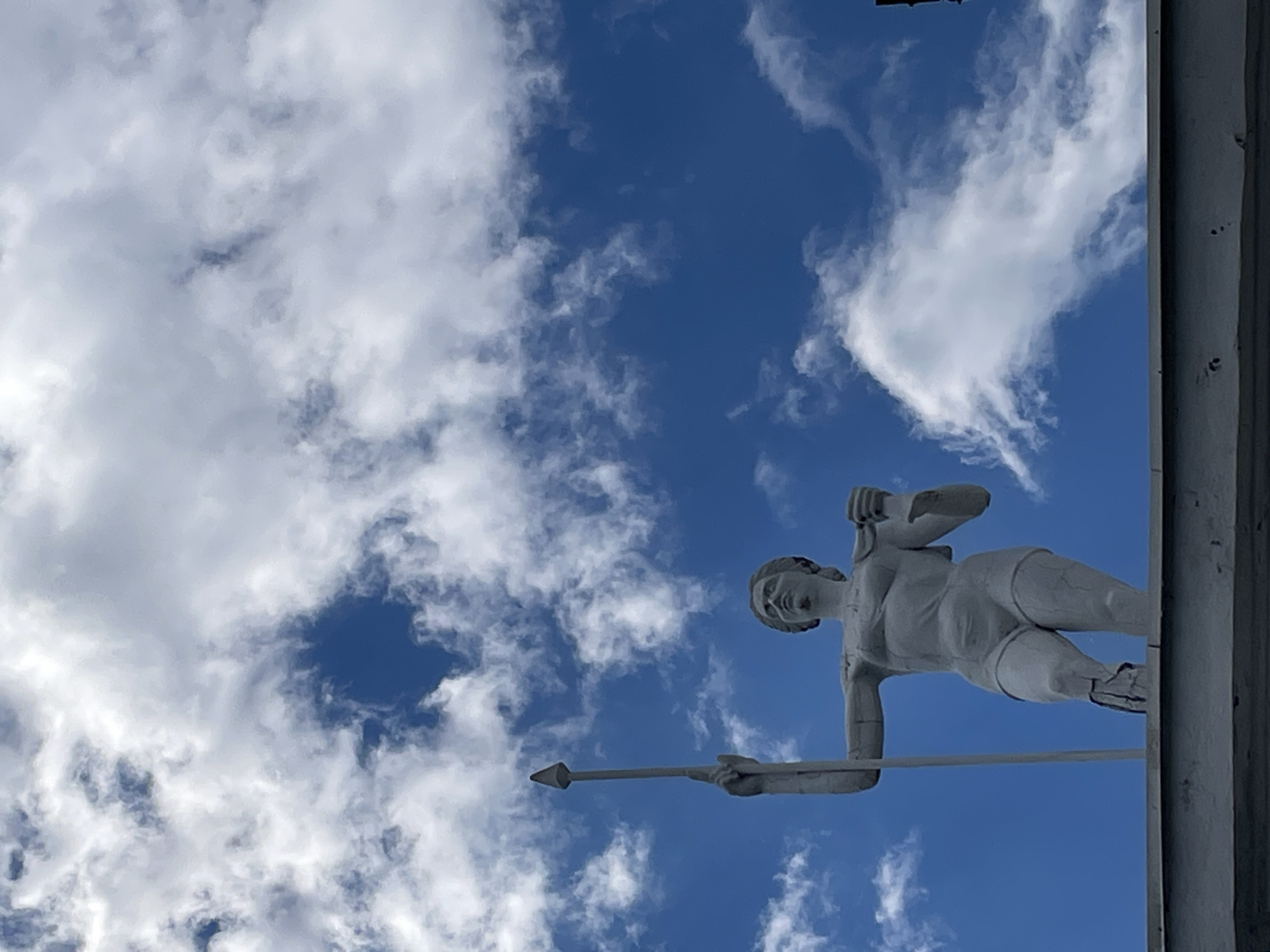 A low-angle, full-shot photograph captures a statue of a woman holding a spear against a backdrop of a bright blue sky dotted with white, puffy clouds. The statue, made of a light-colored material, is positioned on the edge of a grey, horizontal ledge, likely a building rooftop or balcony. The woman is depicted with muscular legs and arms, wearing what appears to be athletic shorts. Her left arm is raised, holding a long, pointed spear upwards, while her right arm is bent at the elbow, with her hand holding an object near her head. The statue's surface shows some wear and tear, with visible cracks on its lower right leg. The lighting suggests it is daytime. The image appears to be taken outdoors in an urban setting, though the specific location is unknown.
