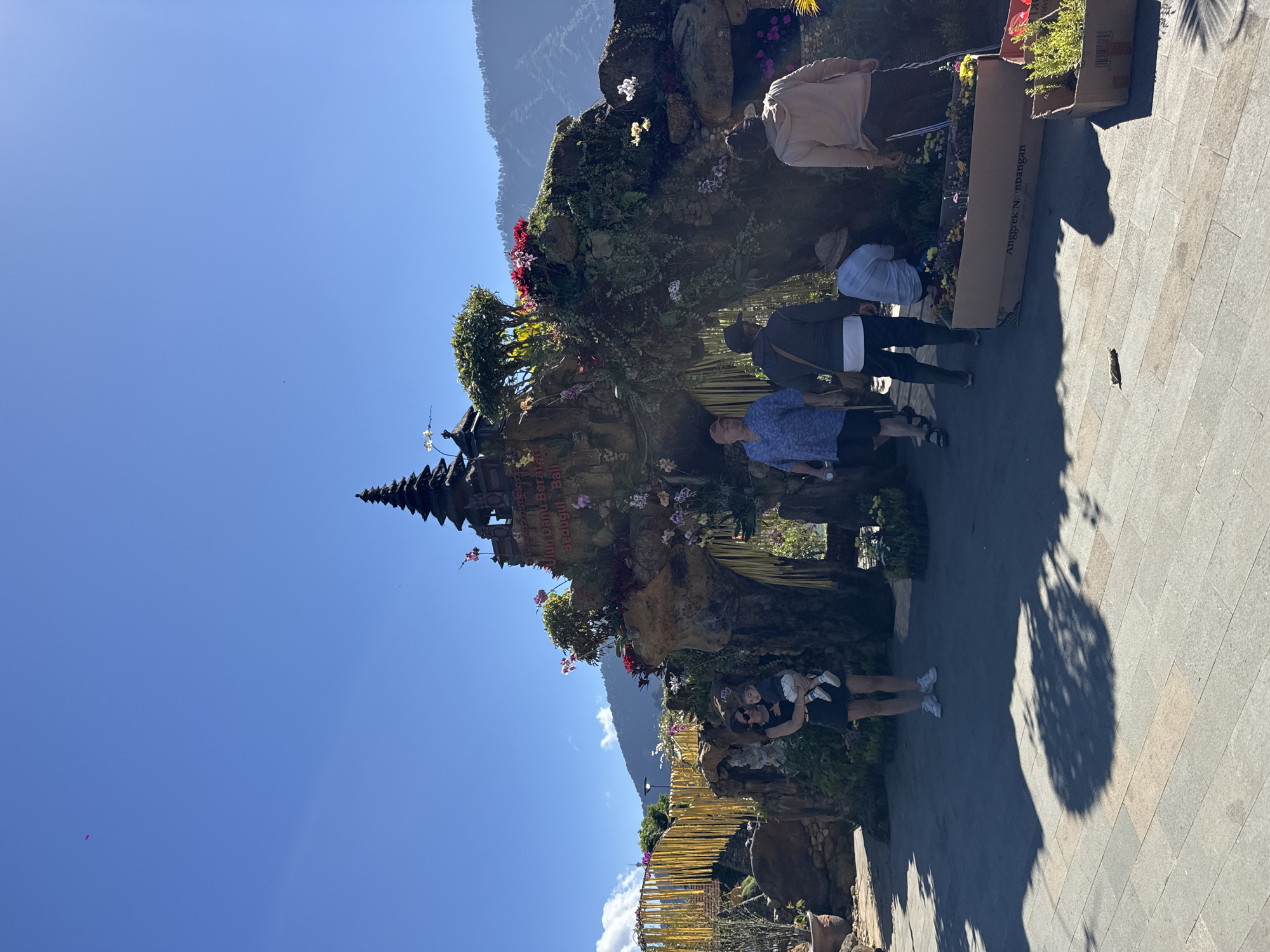 This image depicts the Ulun Danu Beratan Temple in Baturiti, Indonesia, on a clear, sunny day. The foreground features a paved walkway with shadows cast by the sun. Several people are present, including a woman holding a baby, two men walking, and a person kneeling among plants.
The central focus is an elaborately decorated rock structure adorned with lush greenery and vibrant flowers, resembling a temple entrance. A traditional tiered temple roof structure is visible at the top of the rock formation. Behind this, a yellow bamboo-like structure with streamers stretches across the background, possibly a decorative bridge or walkway. In the distance, mountains are visible under a bright blue sky, with a small purple kite also spotted in the sky.
The overall scene suggests a tourist attraction, with people engaged in sightseeing and appreciating the scenic beauty and cultural architecture. The presence of potted plants and decorative elements indicates a well-maintained and picturesque setting.